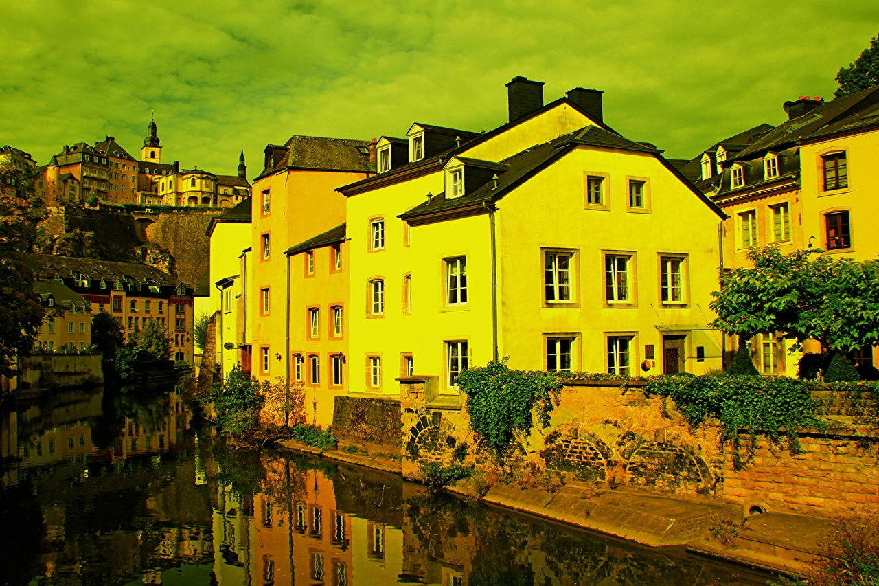 Luxembourg 桌布 壁紙 — 充滿活力的黃色建築物排列在平靜的運河旁，上方是超現實的綠色天空，水面倒影完美。
