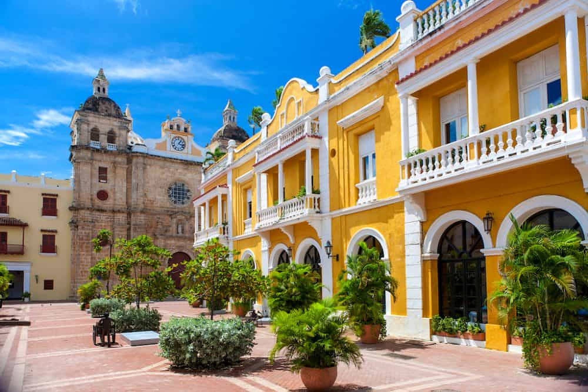 Colombia 桌布 壁紙 — 充滿活力的黃色殖民風格建築，帶有白色陽台，排列在陽光普照的廣場上，旁邊是一座歷史悠久的石砌教堂，上方是清澈的藍天。