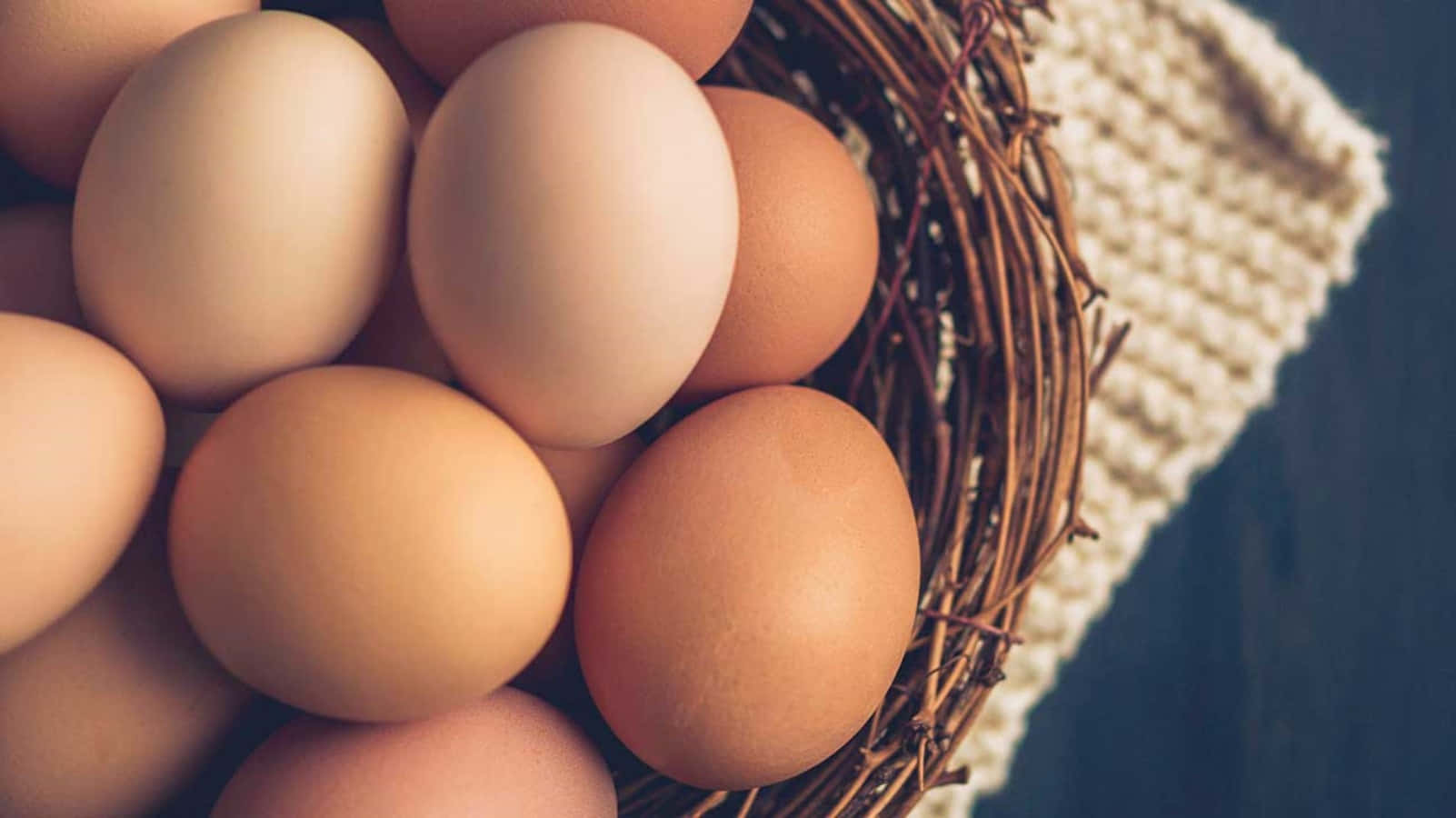 Egg 桌布 壁紙 — 光滑棕色雞蛋的特寫，安放在一個質樸的編織籃子裡，旁邊有一塊奶油色針織布。