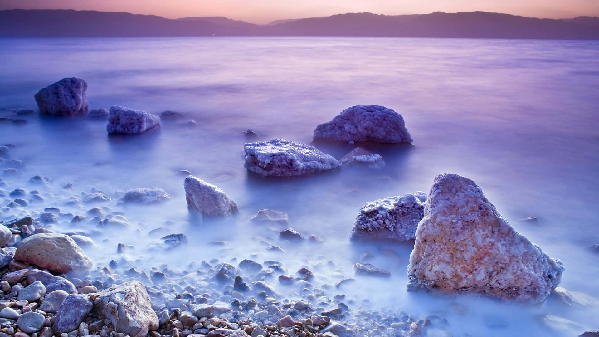 rocky 桌布 壁紙 — 光滑的紫色水流過紋理岩石，在暮色中寧靜的海灘上，遠山相伴。
