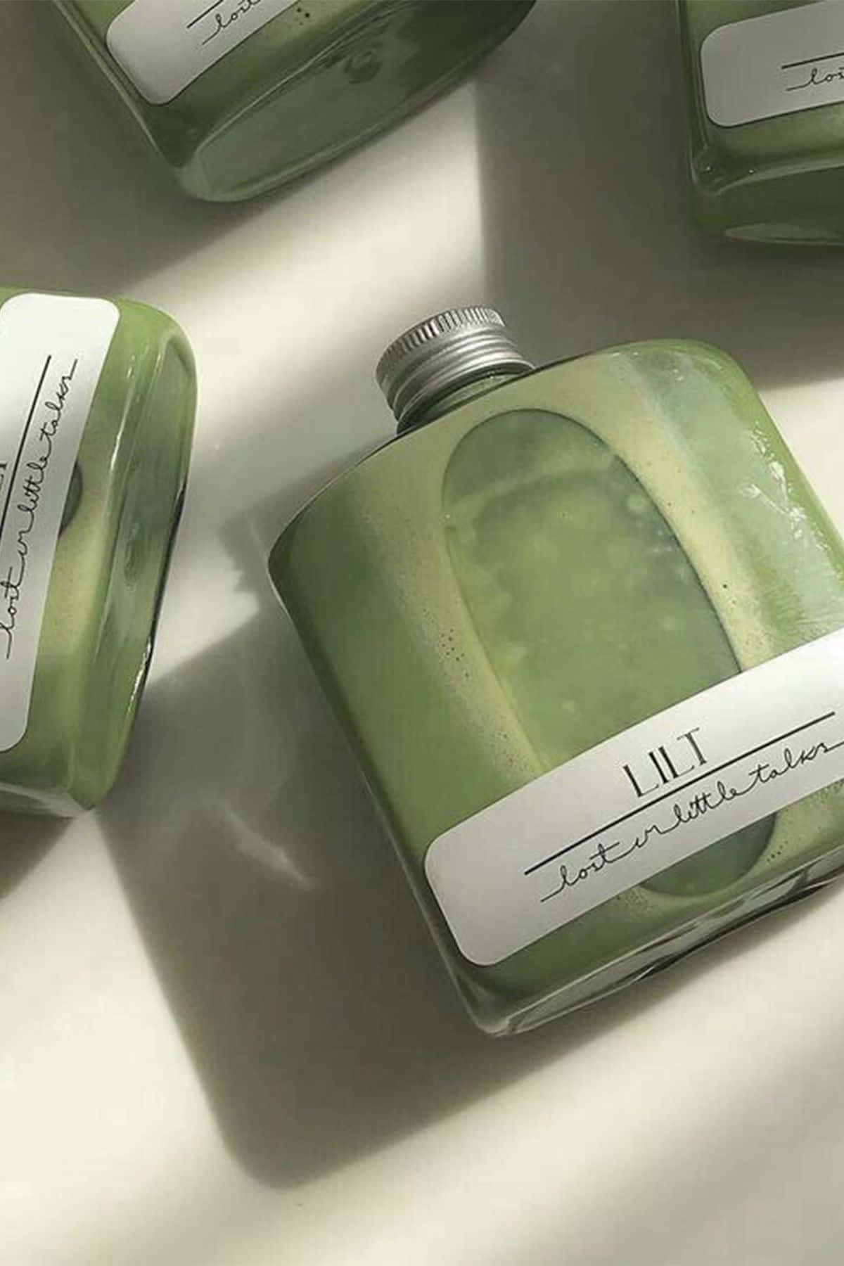 Aesthetic Sage Green 桌布 壁紙 — 光滑的綠色玻璃瓶帶有手寫標籤，在柔和自然光下放置在明亮的白色表面上。