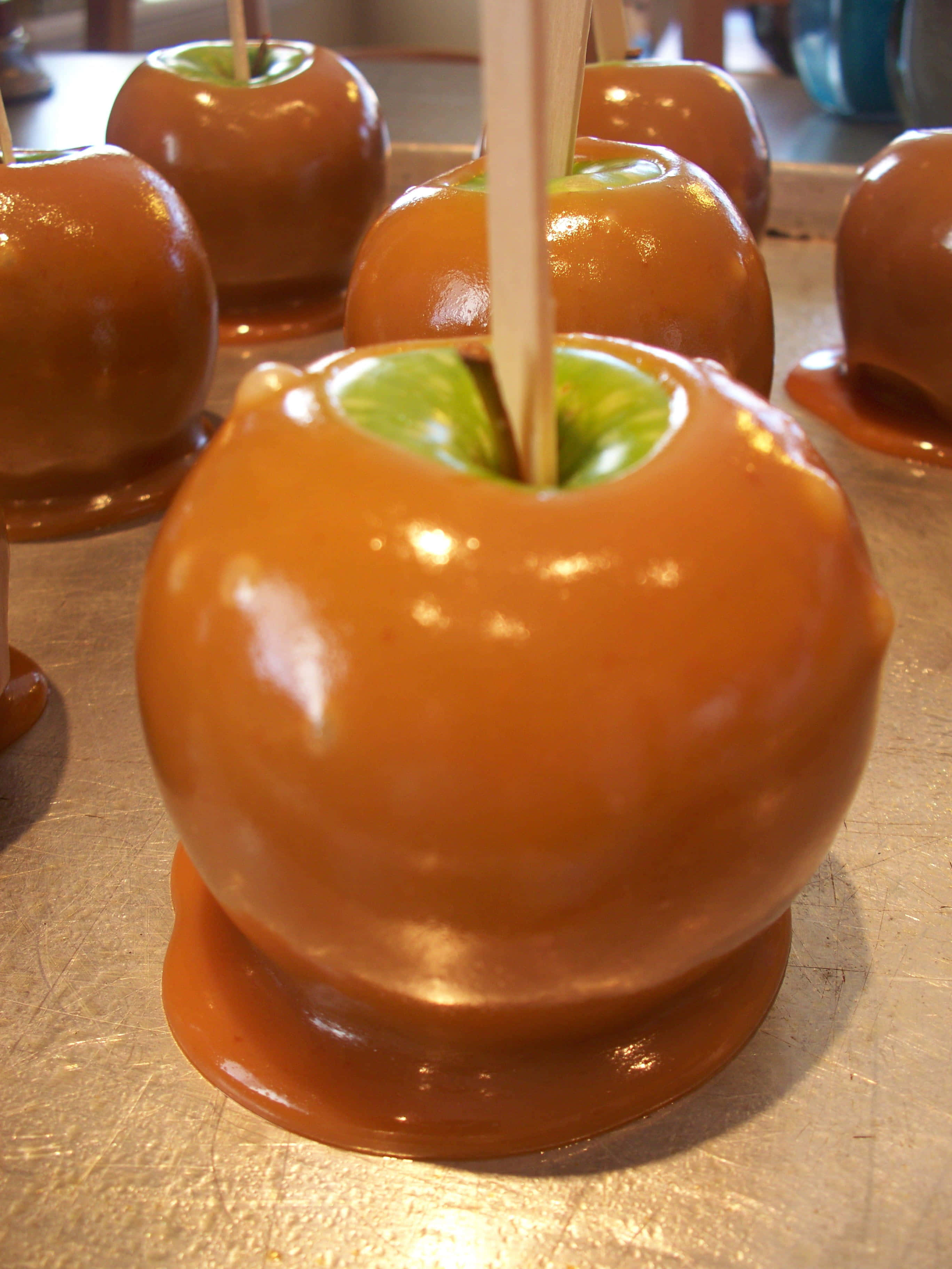 Caramel Apples 桌布 壁紙 — 光澤的焦糖蘋果，帶有綠色莖和木棍，放在金屬托盤上，在溫暖燈光下閃閃發光。