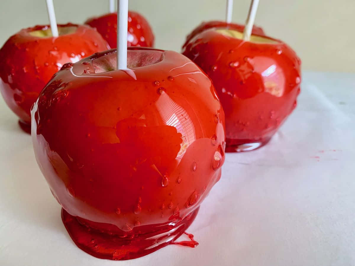 Candy Apples 桌布 壁紙 — 光澤的紅色焦糖蘋果，帶有滴落的焦糖塗層，放在烘焙紙上，插有白色棒子。