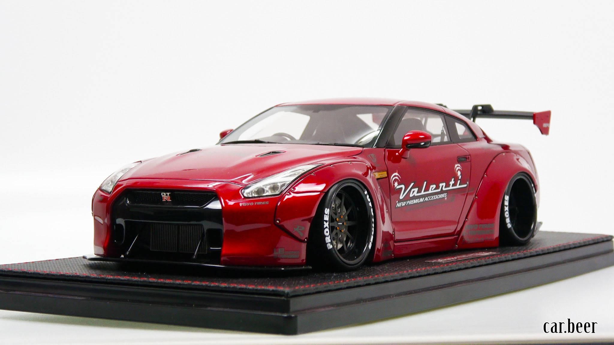 Nissan 桌布 壁紙 — 光澤紅色日產GT-R模型車，配備寬體套件和Valenti貼紙，展示在黑色紋理支架上，背景為白色。
