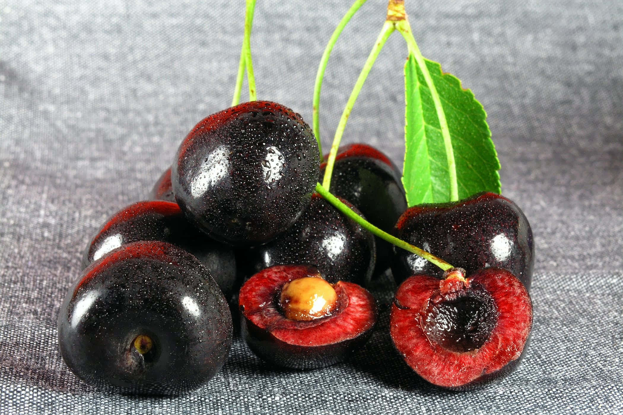 Black Cherry 桌布 壁紙 — 光澤黑色櫻桃帶有鮮紅色內部及綠色莖部，放在有紋理的灰色布料上。