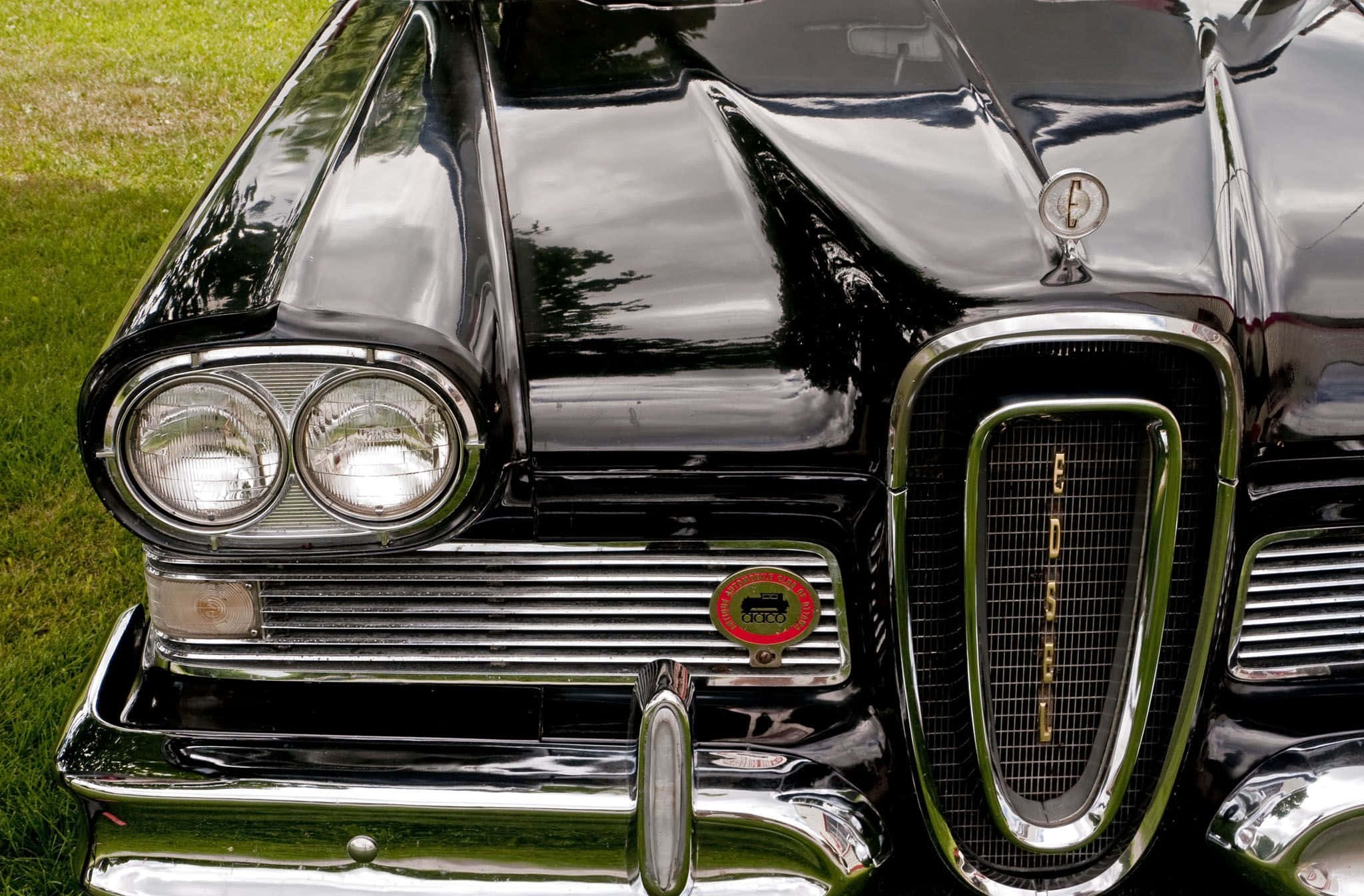 Car Grill 桌布 壁紙 — 光澤黑色經典汽車的前格柵和頭燈特寫，反射出綠色草地和樹木。