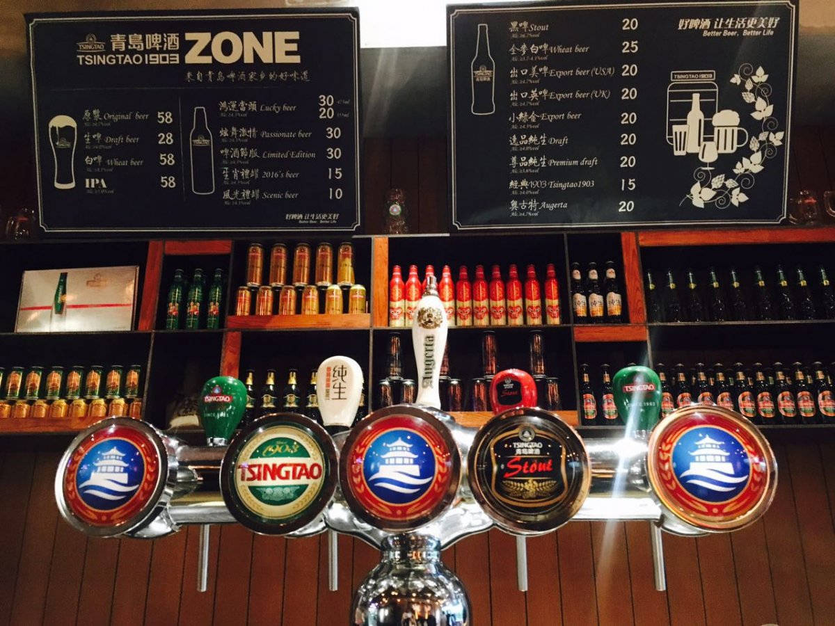 Beer 桌布 壁紙 — 光線充足的吧台龍頭系統，設有青島啤酒龍頭和擺滿色彩繽紛瓶子的架子，下方是黑板菜單。