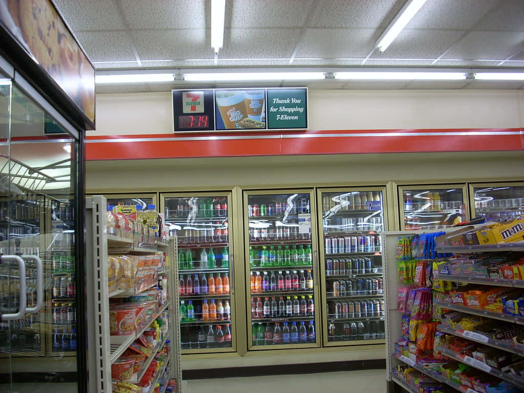 7-ELEVEN 桌布 壁紙 — 光線明亮的7-Eleven便利商店內部，設有螢光燈下的冷藏飲品櫃和零食貨架。