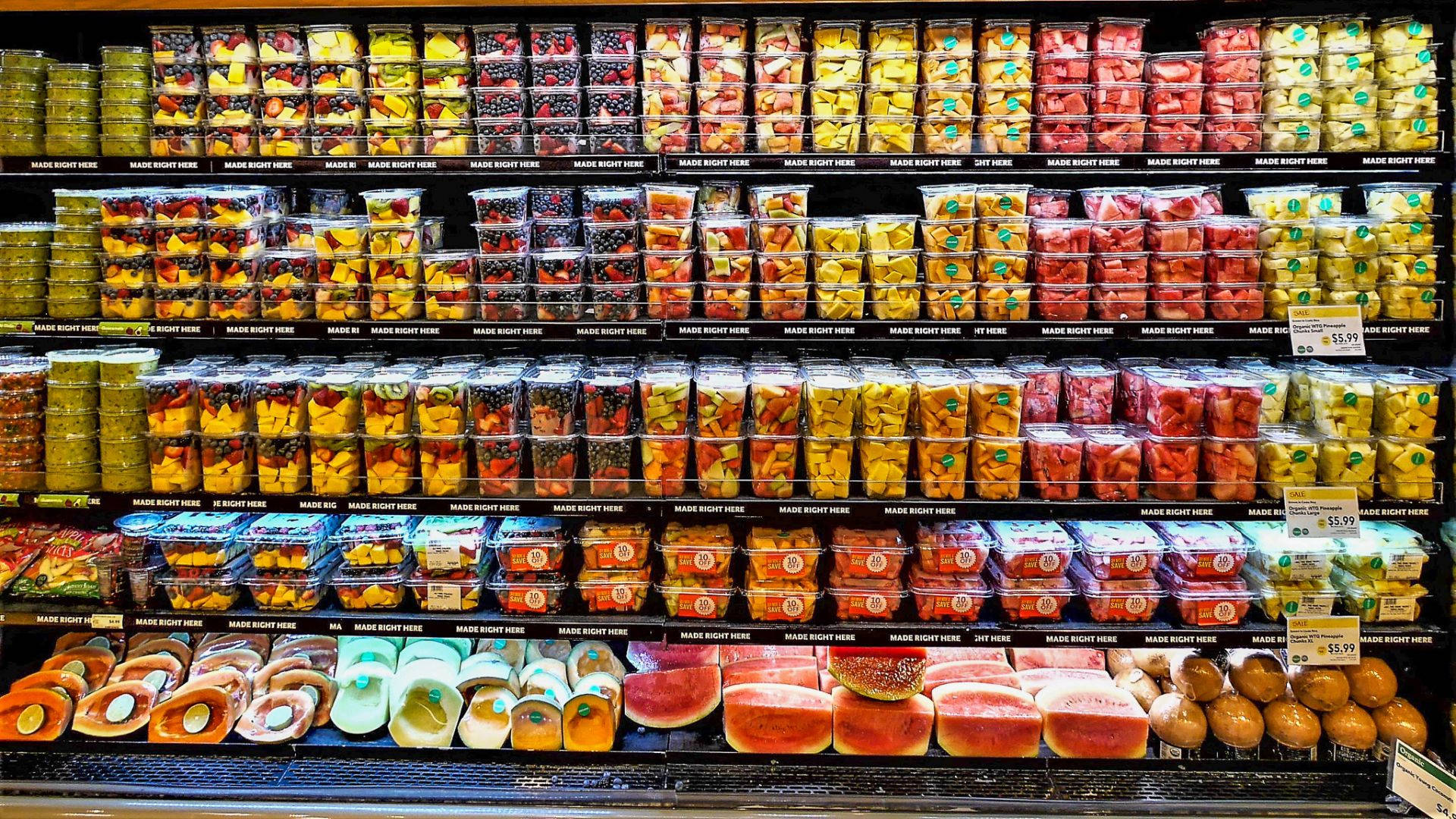 Fruits 桌布 壁紙 — 光線明亮的雜貨店通道展示整齊排列的貨架，上面擺放色彩繽紛的預切水果和蔬菜包裝。
