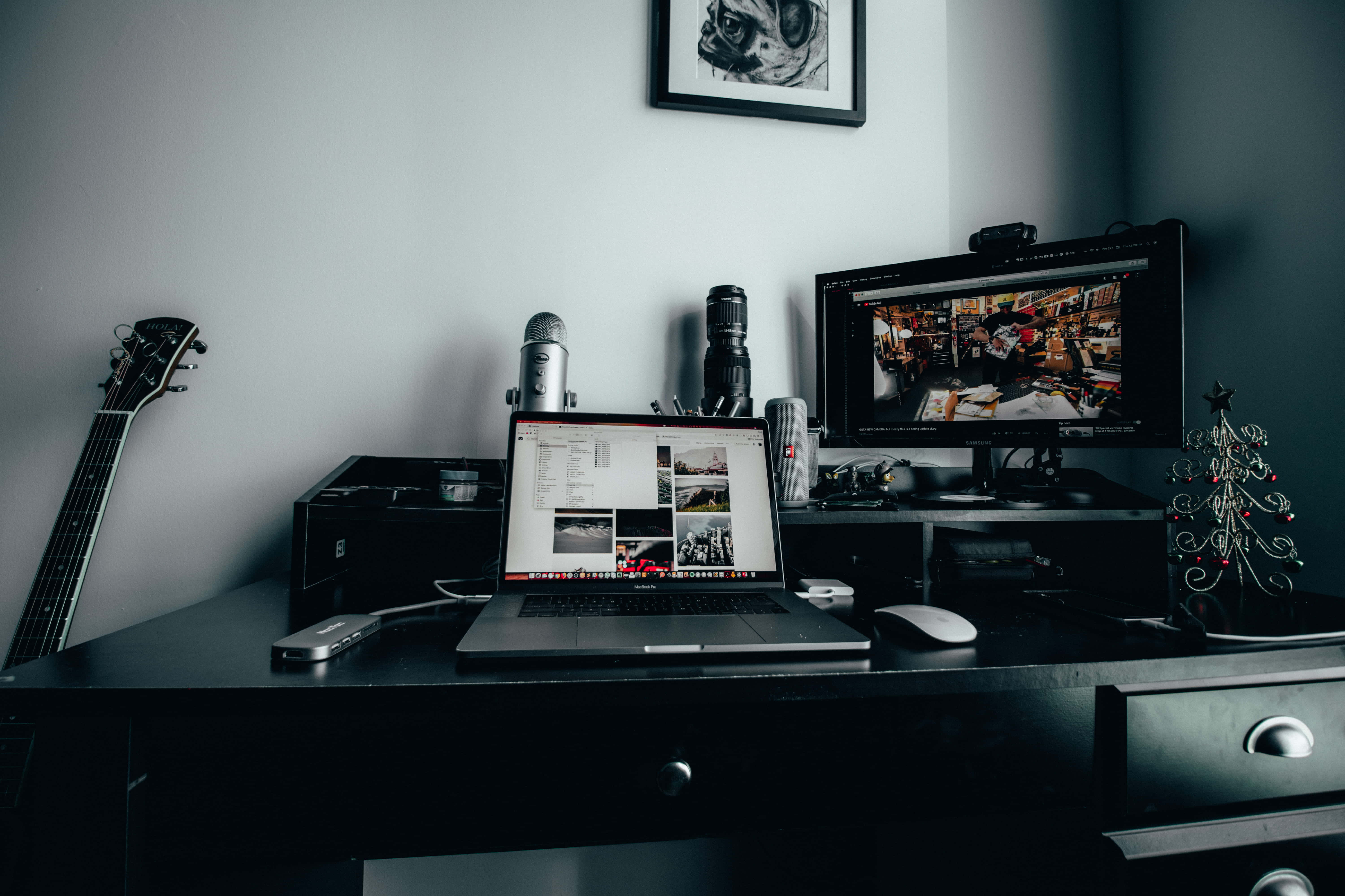 Home Entertainment 桌布 壁紙 — 光線昏暗的家庭辦公室書桌，配有筆記型電腦、相機、顯示器、吉他和裱框的狗肖像，營造出創意工作空間的氛圍。