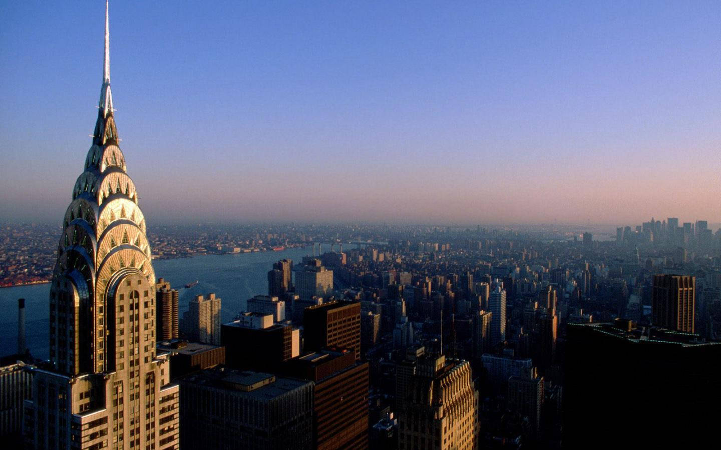 Skyscraper 桌布 壁紙 — 克萊斯勒大廈在黎明時分聳立在紐約市廣闊的天際線上，金色的光芒沐浴著它的裝飾藝術尖頂和周圍的摩天大樓。