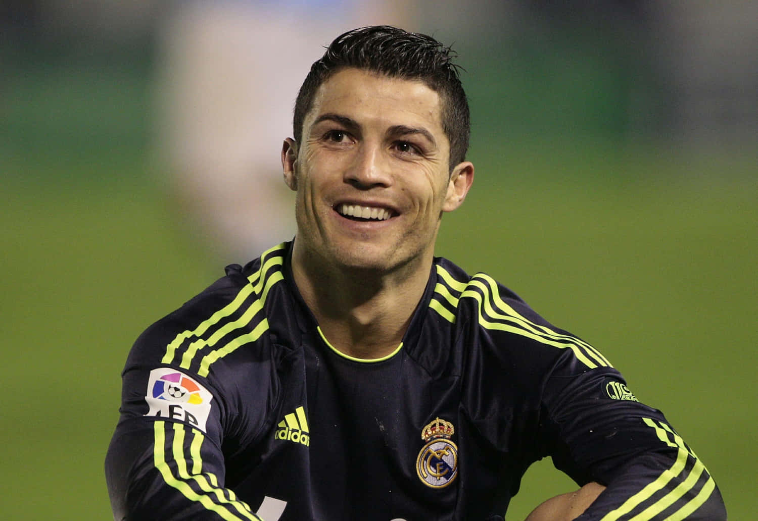 Ronaldo 桌布 壁紙 — 克里斯蒂亞諾·羅納度在模糊的綠色球場背景下，穿著深藍色皇家馬德里訓練衫搭配螢光黃色條紋，燦爛微笑。