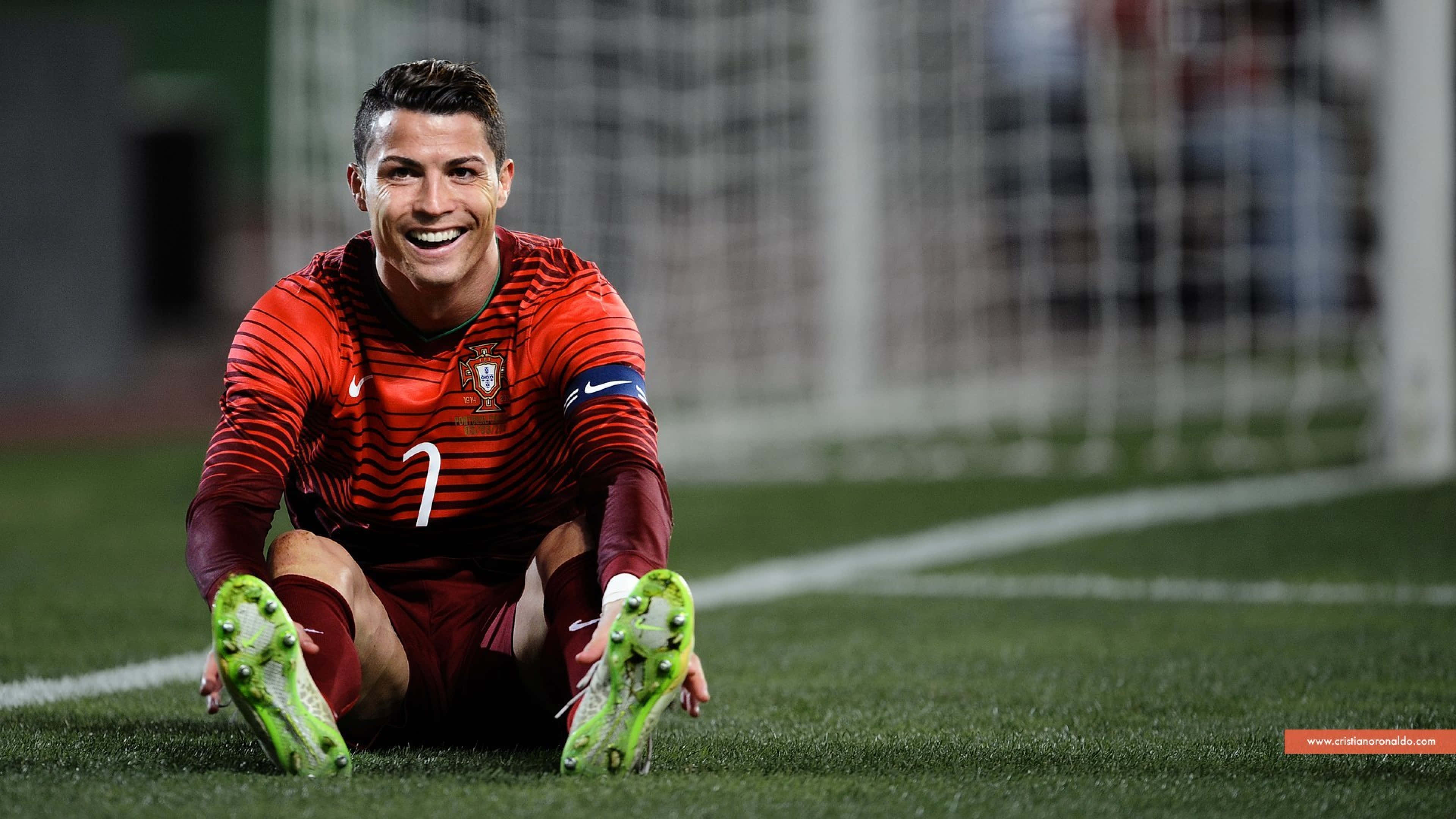 Cristiano Ronaldo 足球 桌布 壁紙 — 克里斯蒂亞諾·羅納度坐在綠色足球場上，身穿紅色葡萄牙球衣和綠色球鞋，露出燦爛笑容。