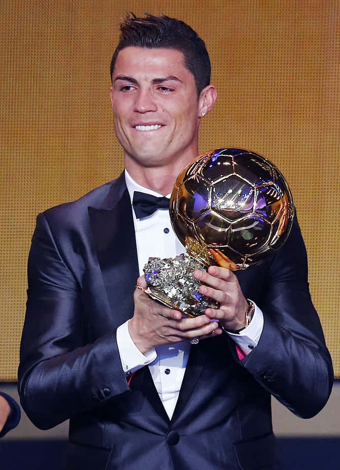 Ronaldo 桌布 壁紙 — 克里斯蒂亞諾·羅納度穿著正式燕尾服，在溫暖燈光背景下自豪地微笑，手持閃閃發光的金色足球獎盃。