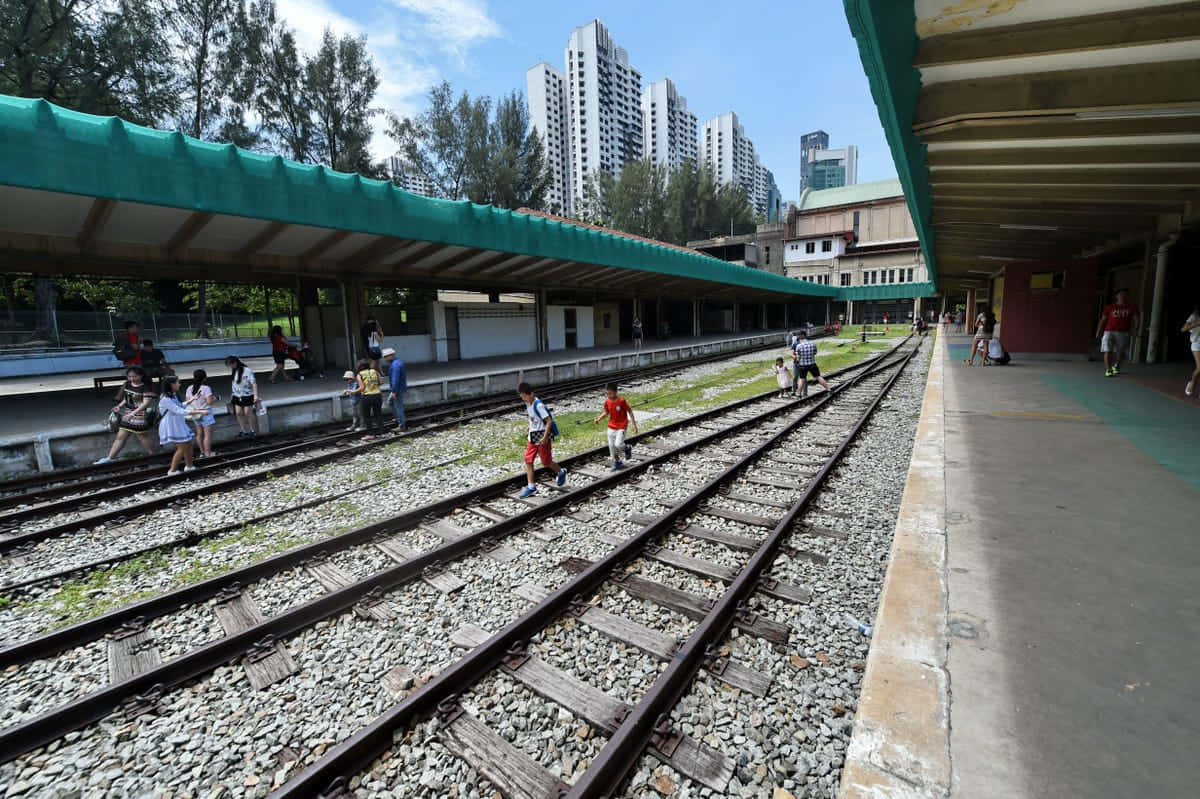 Train Tracks 桌布 壁紙 — 兒童和成人沿著風化的火車軌道行走，上方有綠色天篷，背景可見現代高樓大廈。