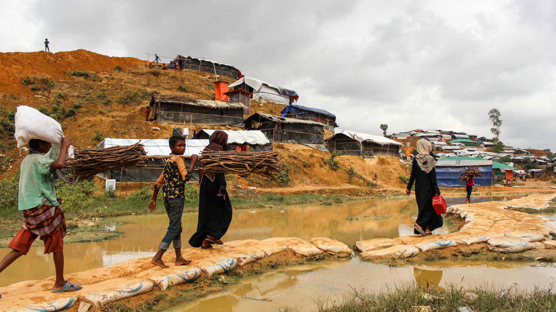 Bangladesh 桌布 壁紙 — 兒童和成人背負重物穿過泥濘水道，在陰天下一片貧瘠山坡上的臨時定居點。