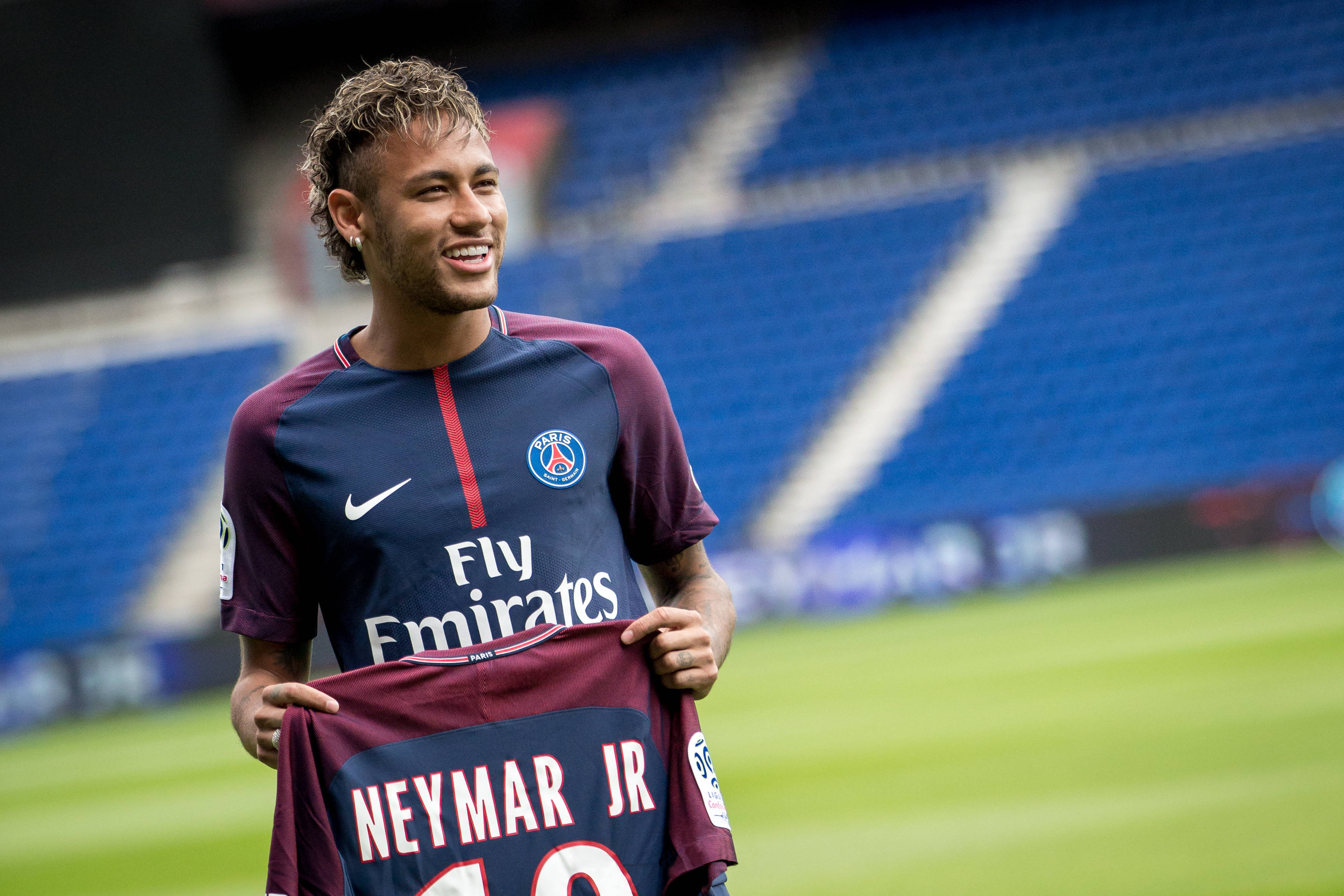 巴西 桌布 壁紙 — 內馬爾（Neymar Jr）在陽光普照的球場上，手持巴黎聖日耳曼球衣，露出燦爛的笑容。