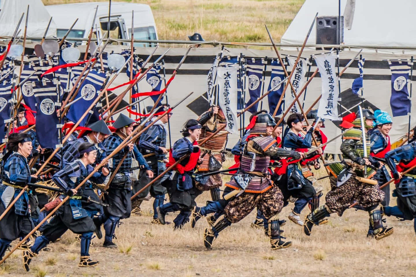 Samurai Battle 桌布 壁紙 — 全副武裝的武士戰士在乾草地上揮舞長矛和旗幟，進行動態的歷史重演。