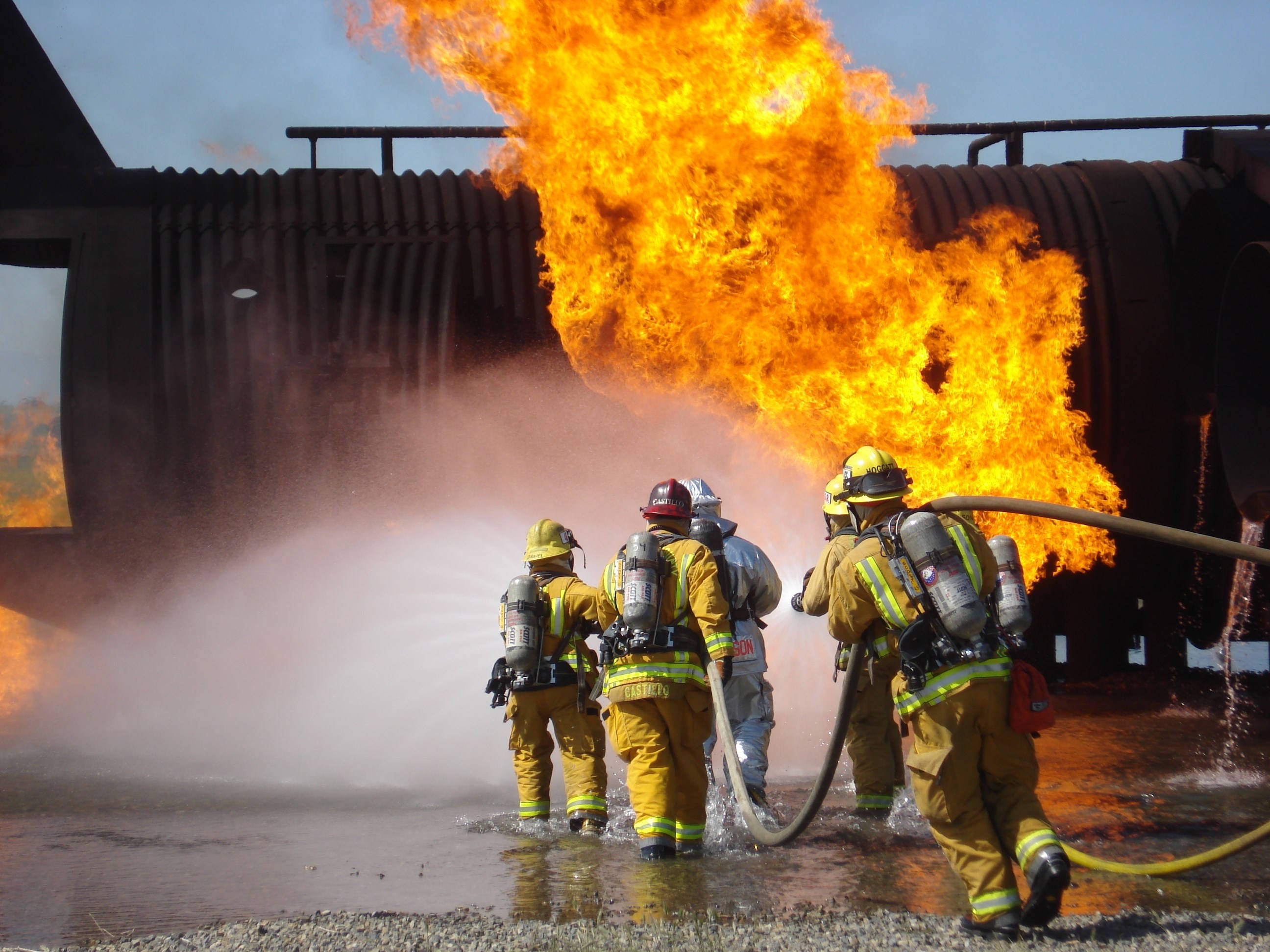 Firefighter 桌布 壁紙 — 全副裝備的消防員在晴朗的藍天下，撲救從大型工業儲罐中噴出的猛烈大火。