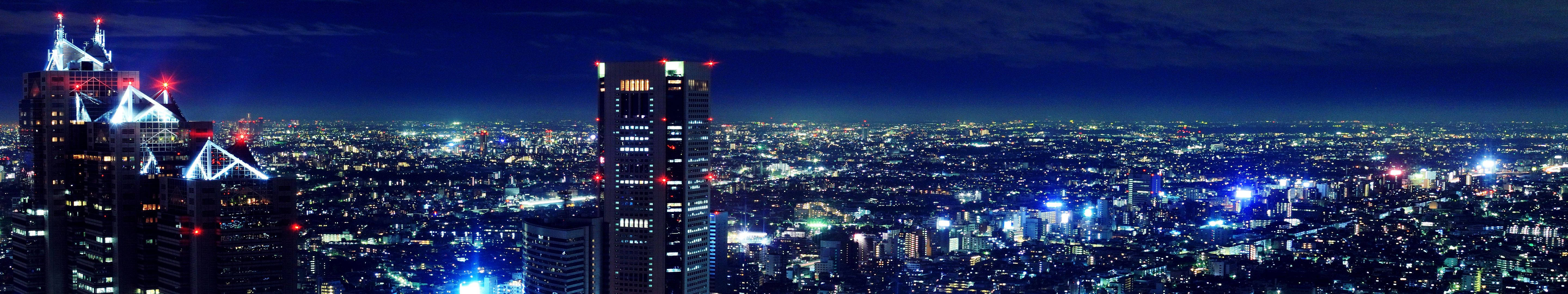 城市風景 桌布 壁紙 — 全景夜景，廣闊的城市景觀中高聳的摩天大樓在黑暗的天空下閃爍著藍色和紅色的燈光。
