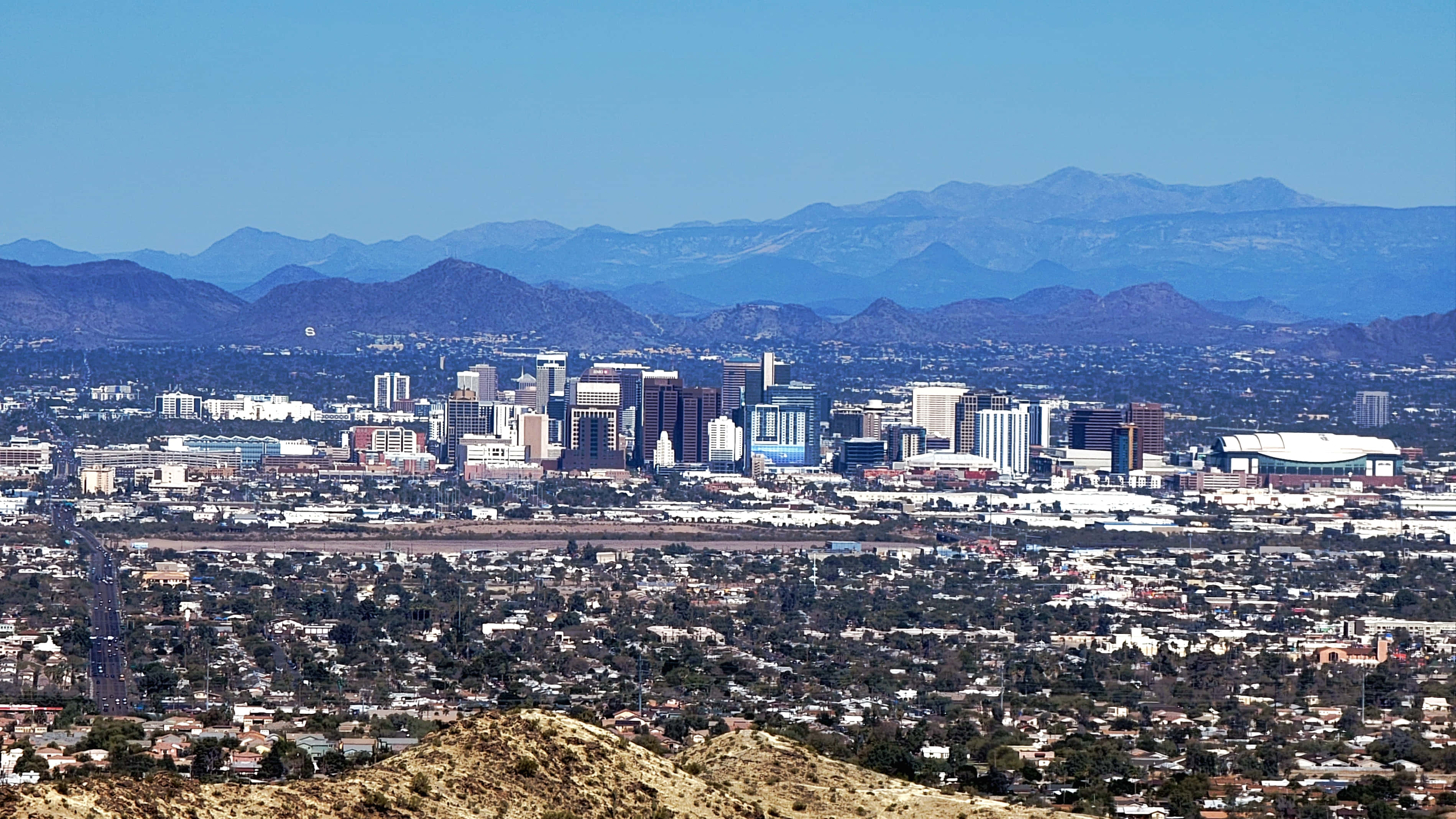 Phoenix Arizona 桌布 壁紙 — 全景視野，展現廣闊的城市景觀，現代摩天大樓坐落於住宅區與遠處山脈之間，天空清澈湛藍。