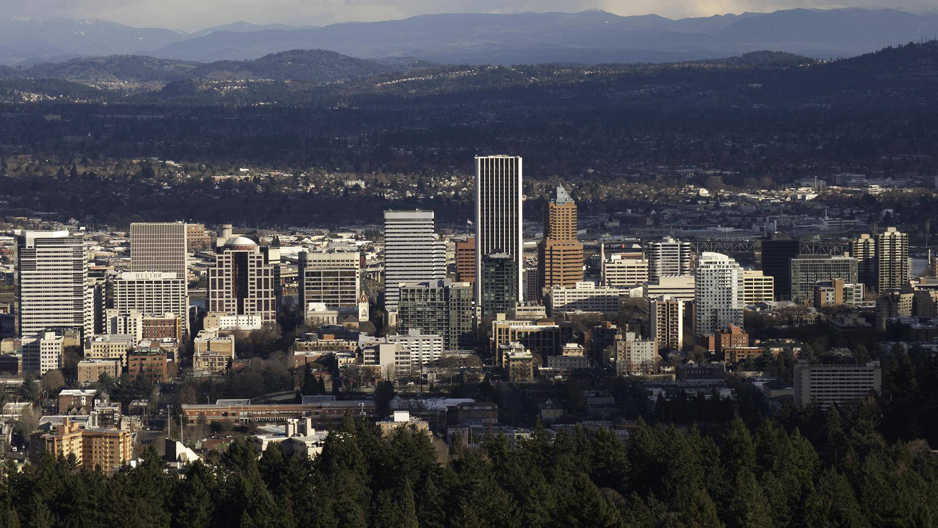 Portland 桌布 壁紙 — 全景視野的城市天際線，現代摩天大樓坐落在茂密森林和遠處山脈之間，天空朦朧。