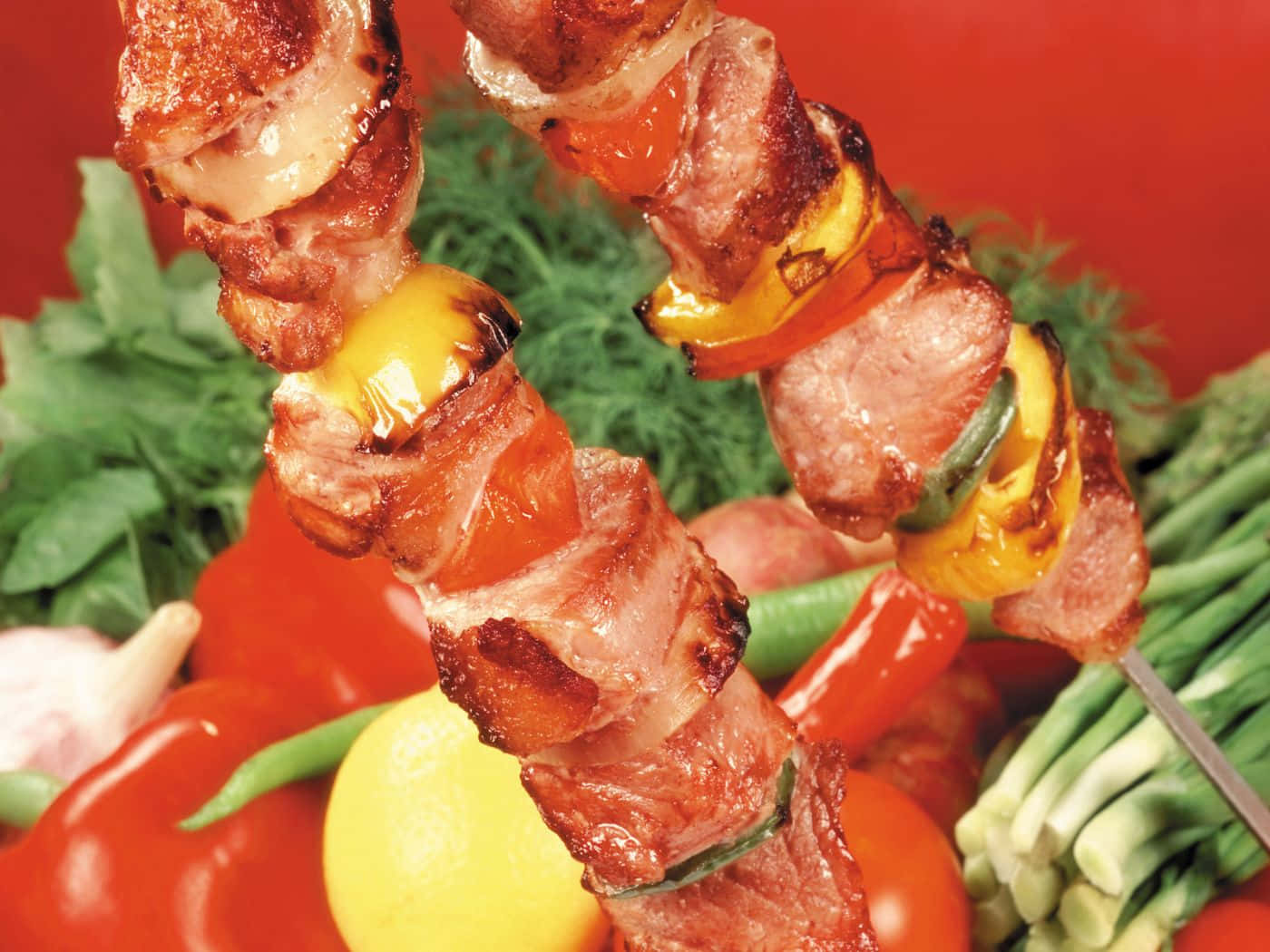 Bbq 桌布 壁紙 — 兩串滋滋作響的烤肉和彩色甜椒放在新鮮蔬菜的鮮豔床上，背景為大膽的紅色。