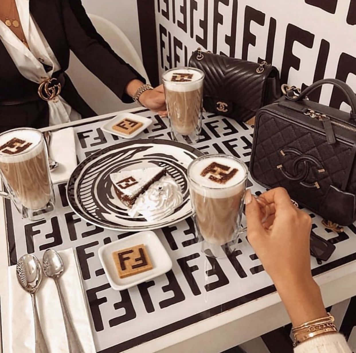Fendi 桌布 壁紙 — 兩人在一張覆蓋著大膽Fendi標誌圖案的桌子上享用豪華咖啡和蛋糕，附近有設計師手袋。