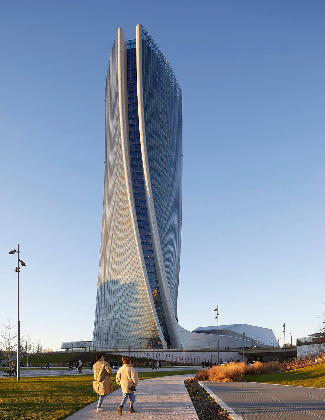 Skyscraper 桌布 壁紙 — 兩人在一條小徑上漫步，上方是宏偉的流線型玻璃摩天大樓，帶有弧形外牆，在清澈的藍天下。