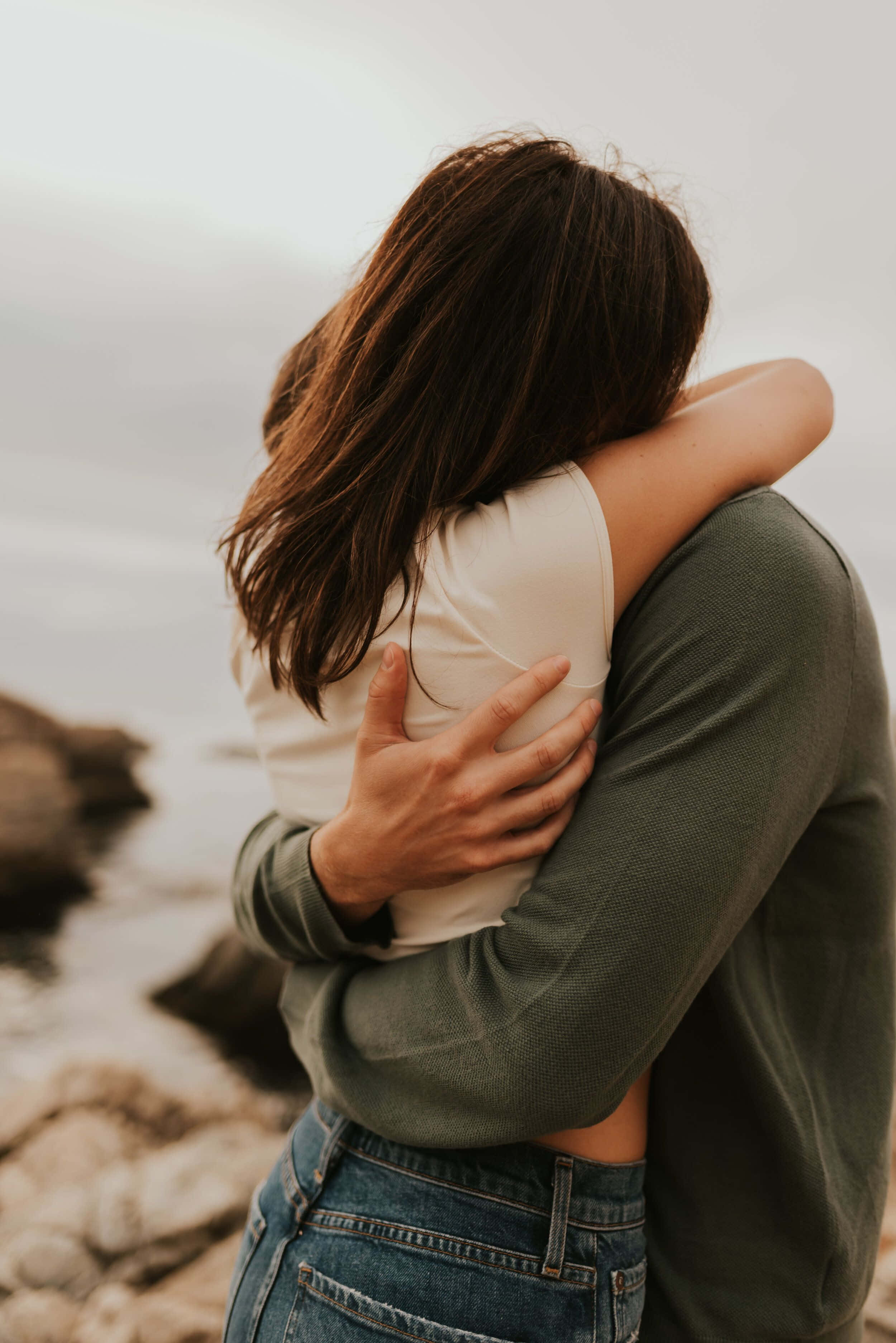 Hugging 桌布 壁紙 — 兩人在多雲的天空下，在岩石海岸線上溫暖地擁抱。
