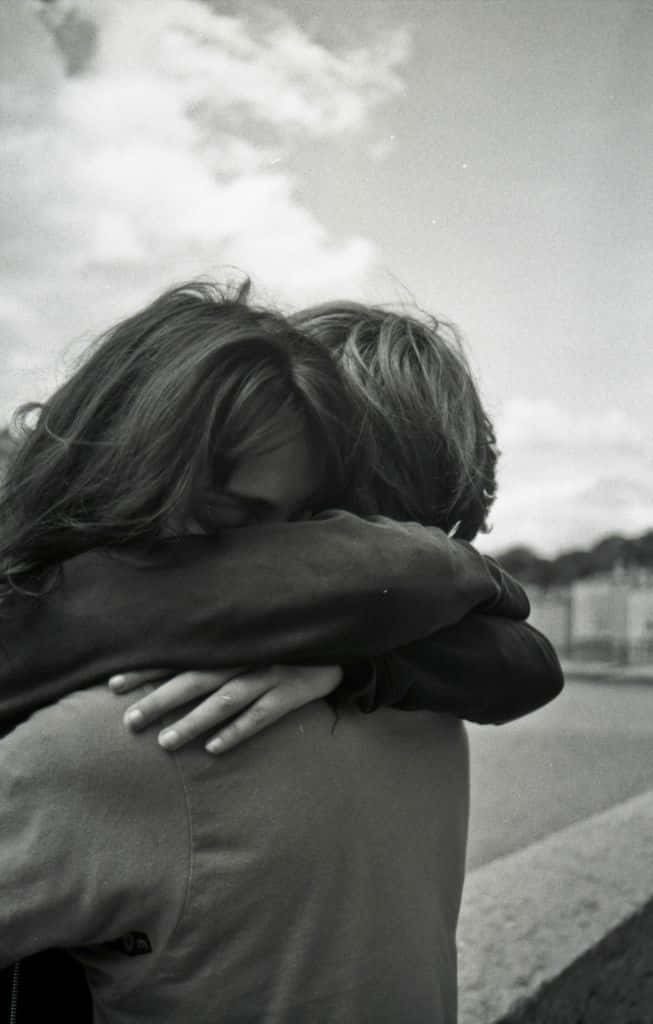 Hugging 桌布 壁紙 — 兩人在多雲的天空下從背後緊緊擁抱，呈現溫柔的黑白瞬間。