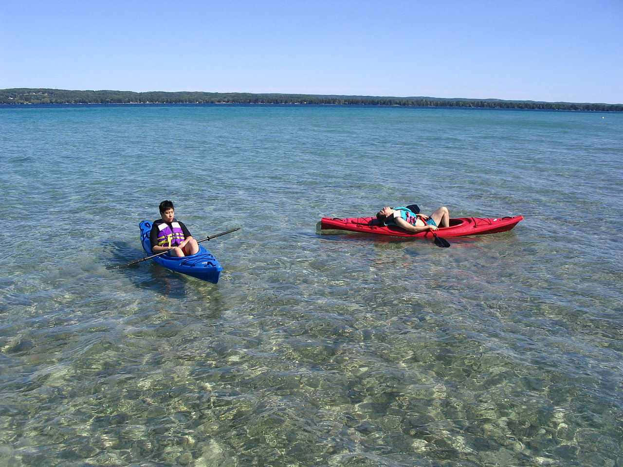 Torch Lake 桌布 壁紙 — 兩人在平靜清澈的湖上划著獨木舟放鬆，頭頂是明亮的藍天，遠處是樹木繁茂的湖岸。