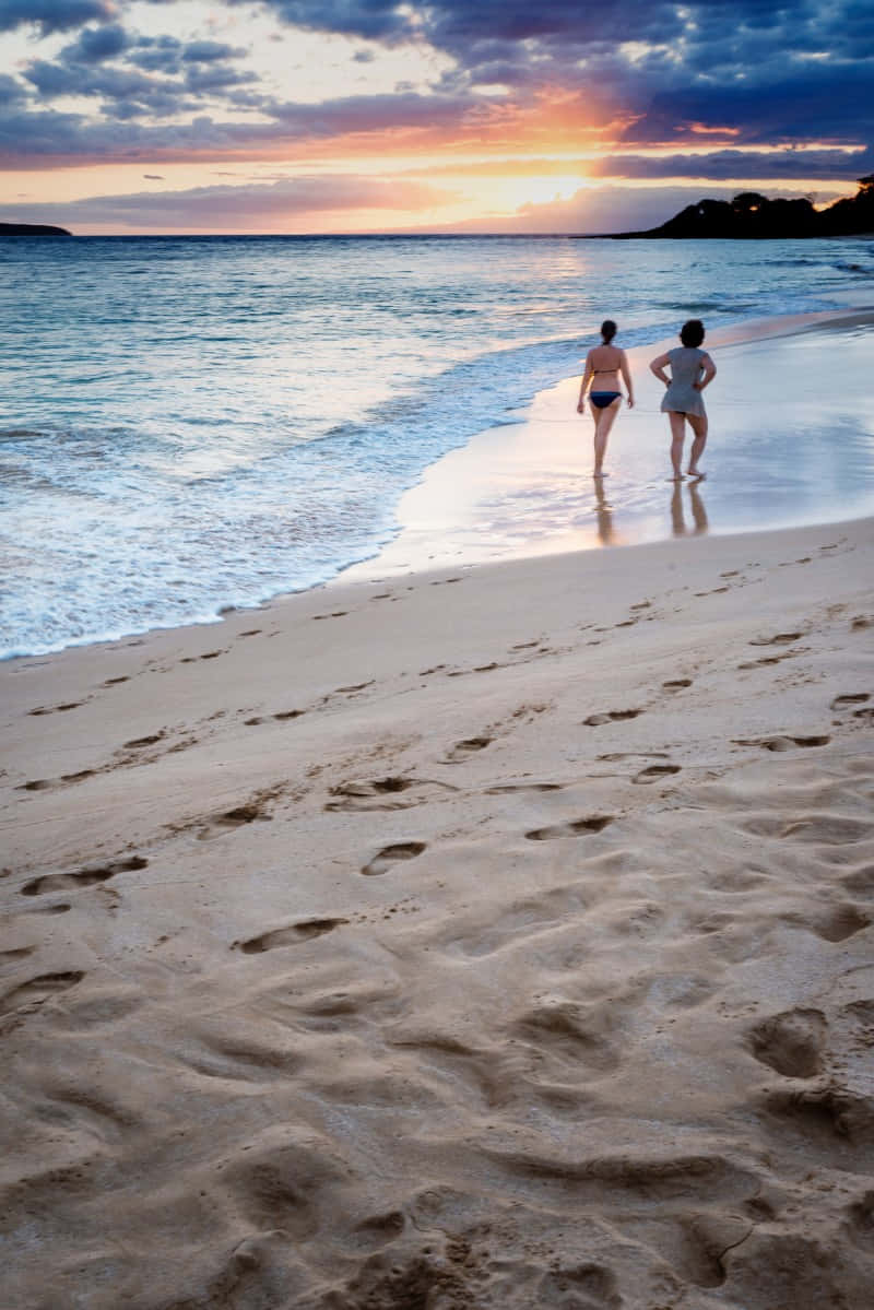 Beach Walk 桌布 壁紙 — 兩人在日落時分沿著沙灘漫步，海浪輕拍他們的腳，天空呈現鮮豔的橙色和紫色。