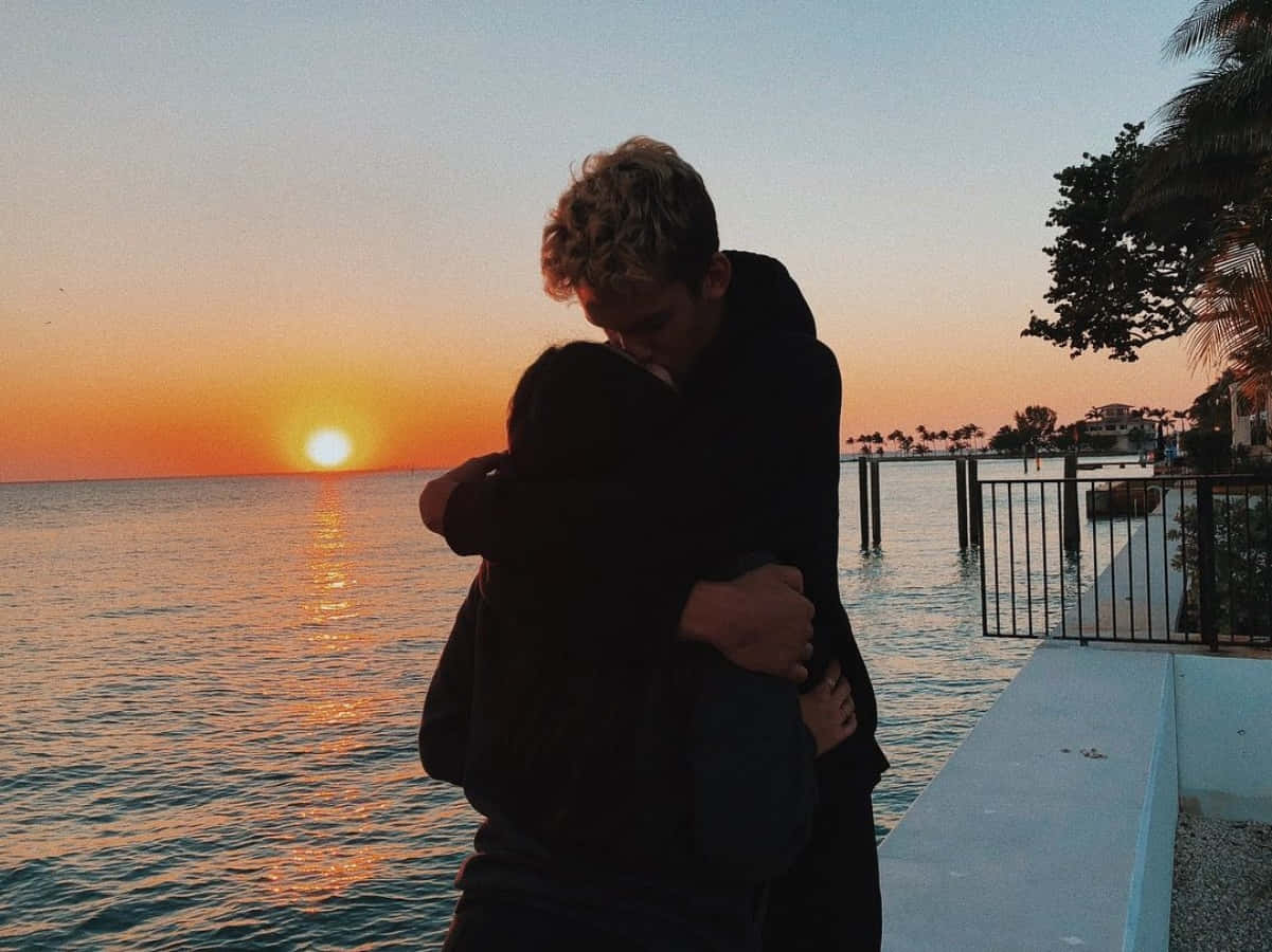 Aesthetic Couple 桌布 壁紙 — 兩人在海邊碼頭溫暖擁抱，夕陽在海面上灑下金色光芒。