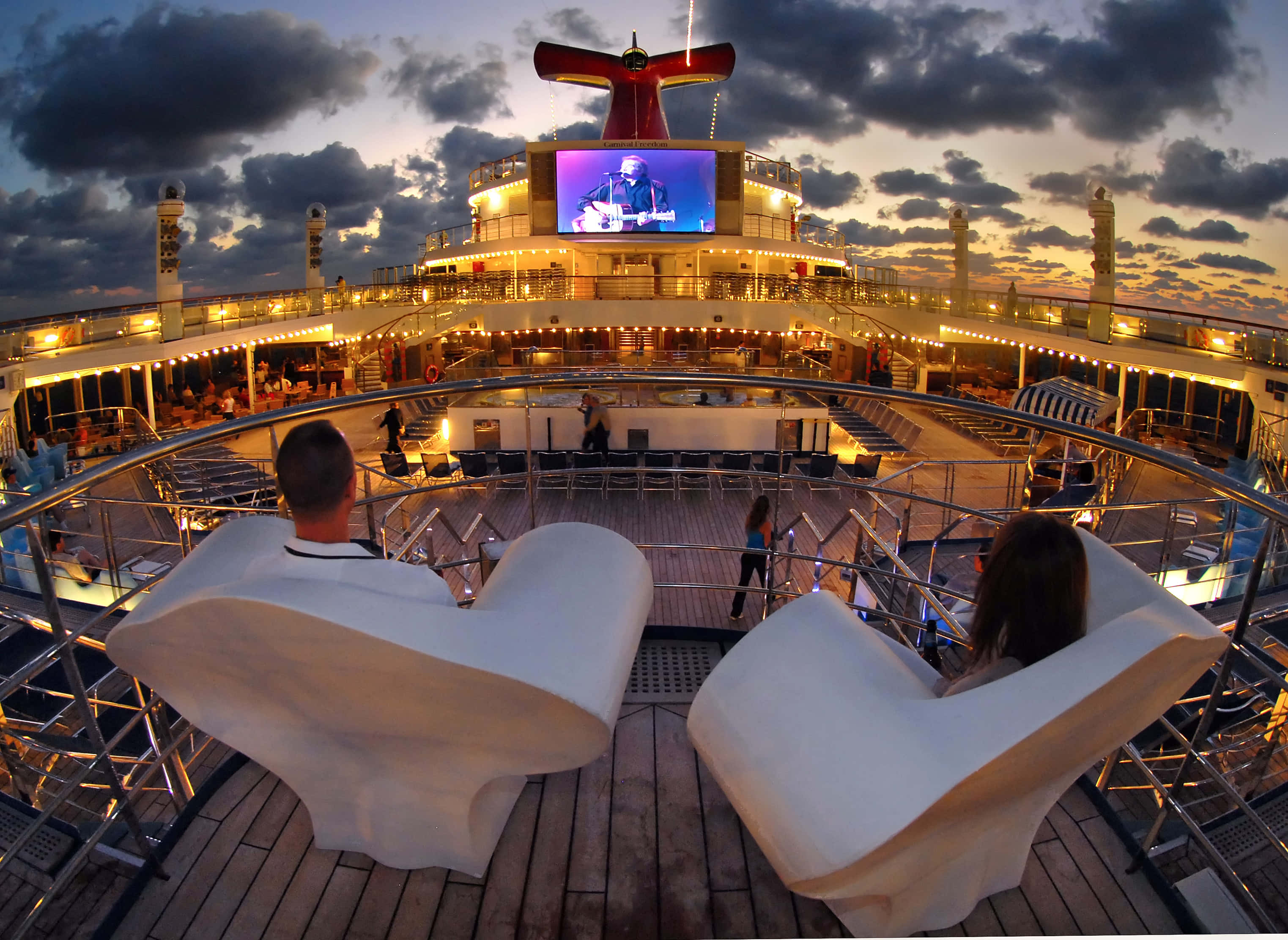 Carnival Conquest 桌布 壁紙 — 兩人在郵輪甲板的白色躺椅上放鬆，一位現場音樂家在暮色天空下表演。