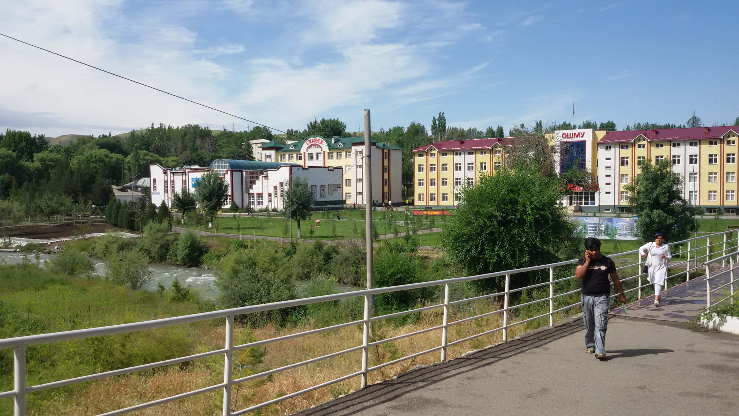 Kyrgyzstan 桌布 壁紙 — 兩人在金屬欄杆橋上漫步，橋下是湍急的河流，背景是色彩繽紛的建築物和茂密的綠樹，頭頂是明亮的藍天。