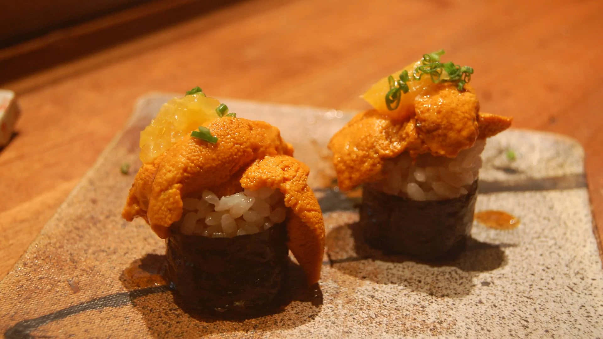 Types Of Sushi 桌布 壁紙 — 兩件鮮橙色海膽壽司配上綠色裝飾，放在斑點盤上，在溫暖燈光下。