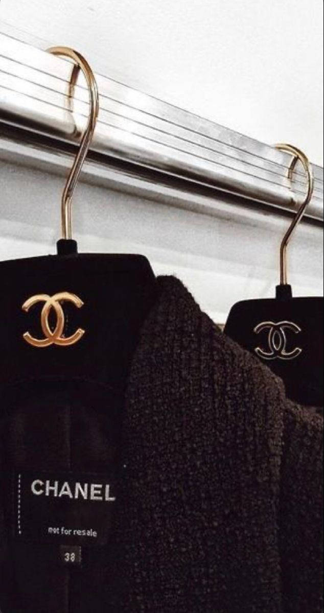 Chanel Aesthetic 桌布 壁紙 — 兩件黑色 Chanel 服裝掛在金色衣架上，掛在帶有可見紋理和品牌標籤的時尚金屬桿上。