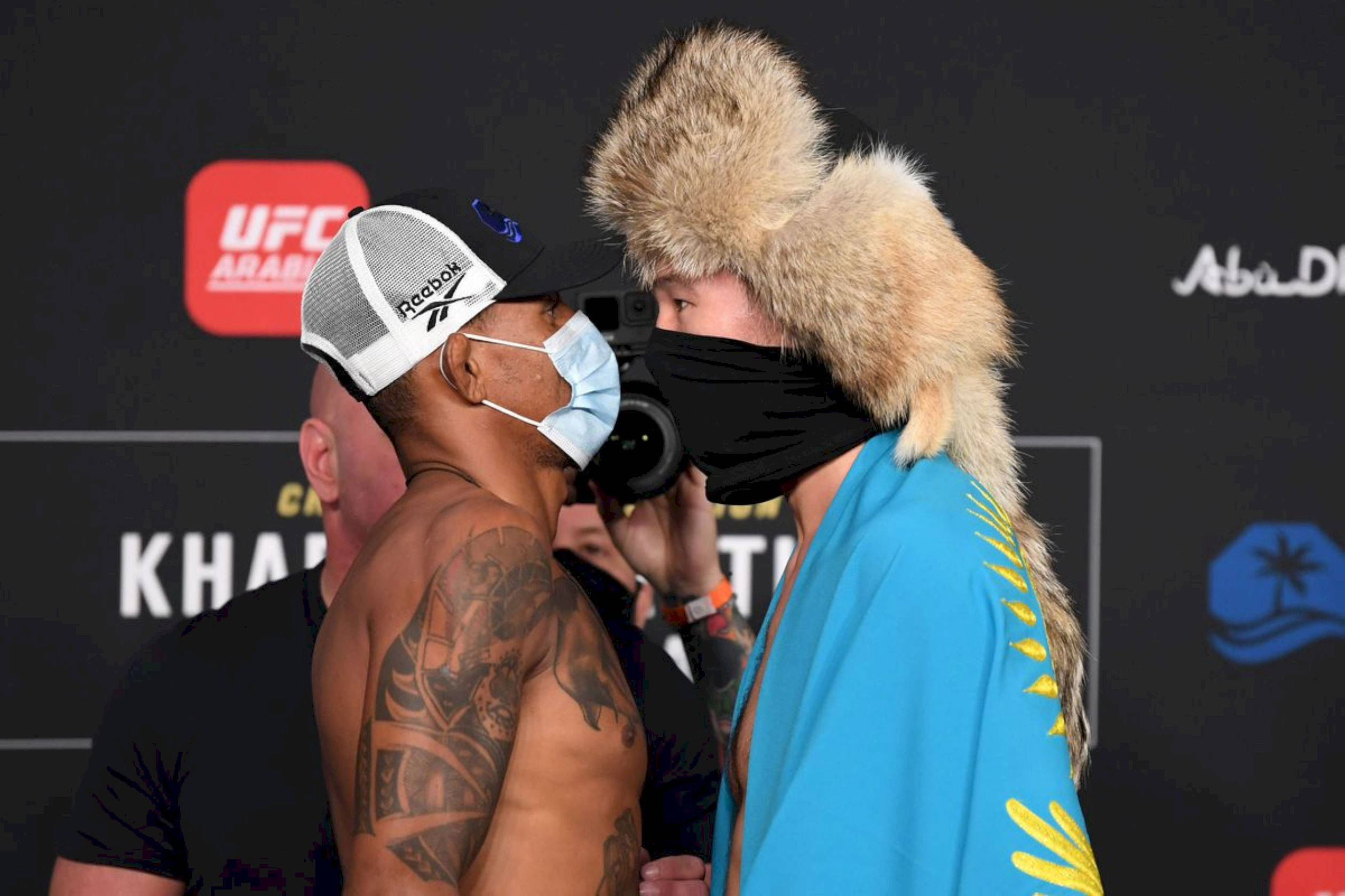 UFC 桌布 壁紙 — 兩位UFC拳手在稱重時對峙，一位戴著毛帽和藍色長袍，另一位戴著帽子和口罩。