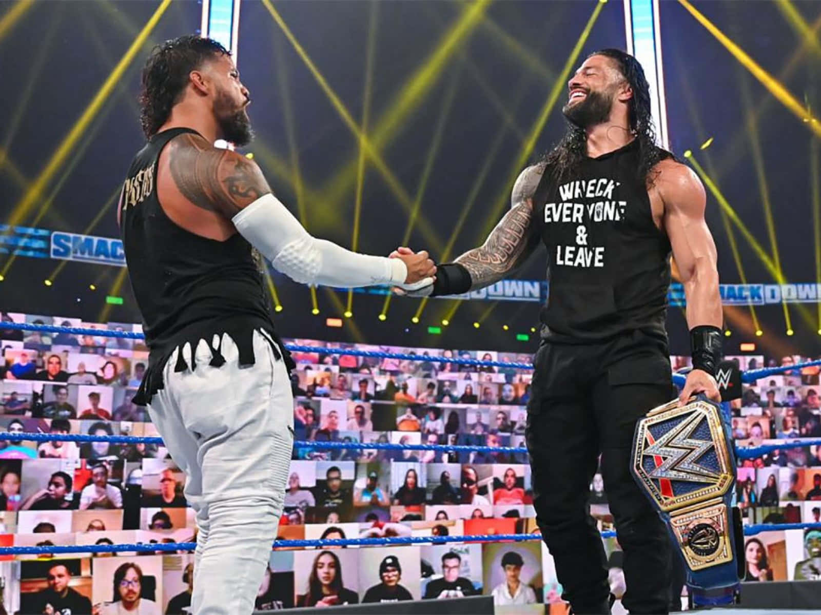 Roman Reigns 桌布 壁紙 — 兩位WWE摔角手在擂台上握手，其中一人手持冠軍腰帶，虛擬粉絲從他們身後的螢幕觀看。