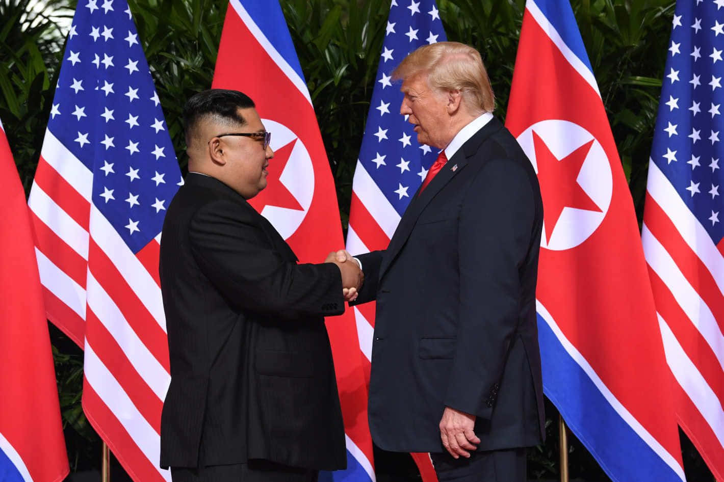 Kim Jong Un 桌布 壁紙 — 兩位世界領袖在交替的美國和北韓國旗前握手，周圍是茂密的綠植。