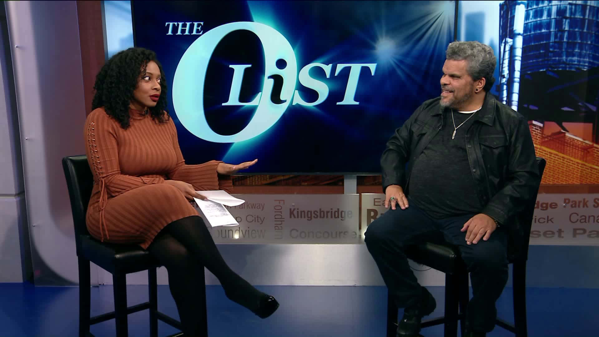 Luis Guzmán 桌布 壁紙 — 兩位主持人坐在黑色凳子上，在一個背後有發光「The List」標誌的攝影棚中交談。