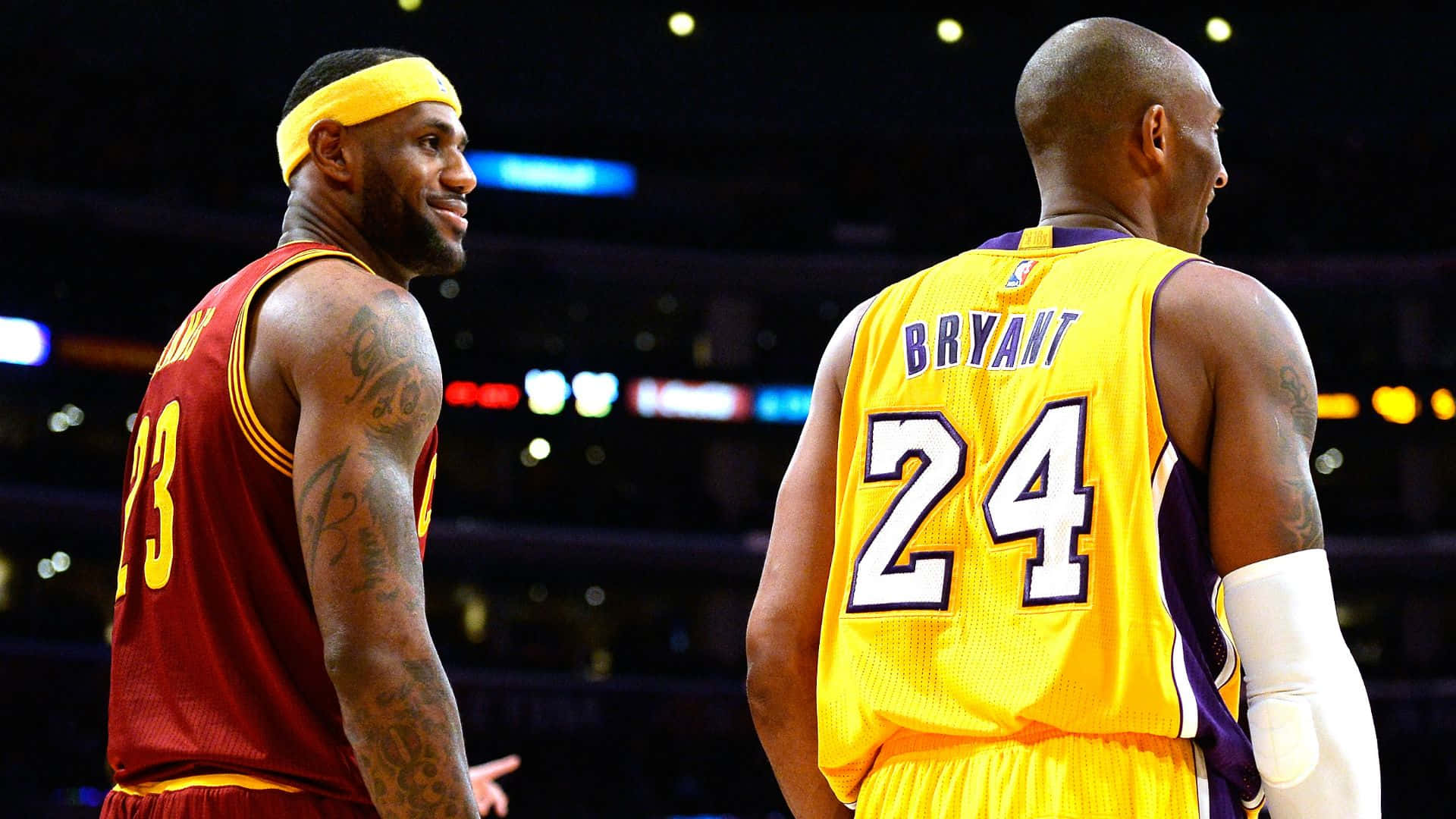 科比· Bryant 桌布 壁紙 — 兩位傳奇籃球運動員穿著鮮豔球衣，在燈光明亮的球場上比賽時面對面。