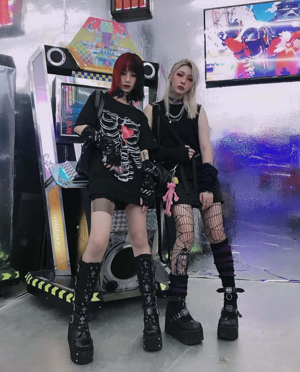 emo 美學 桌布 壁紙 — 兩位哥德少女穿著黑色龐克服裝，在霓虹燈閃爍的遊戲室中，自信地站在發光的街機旁。