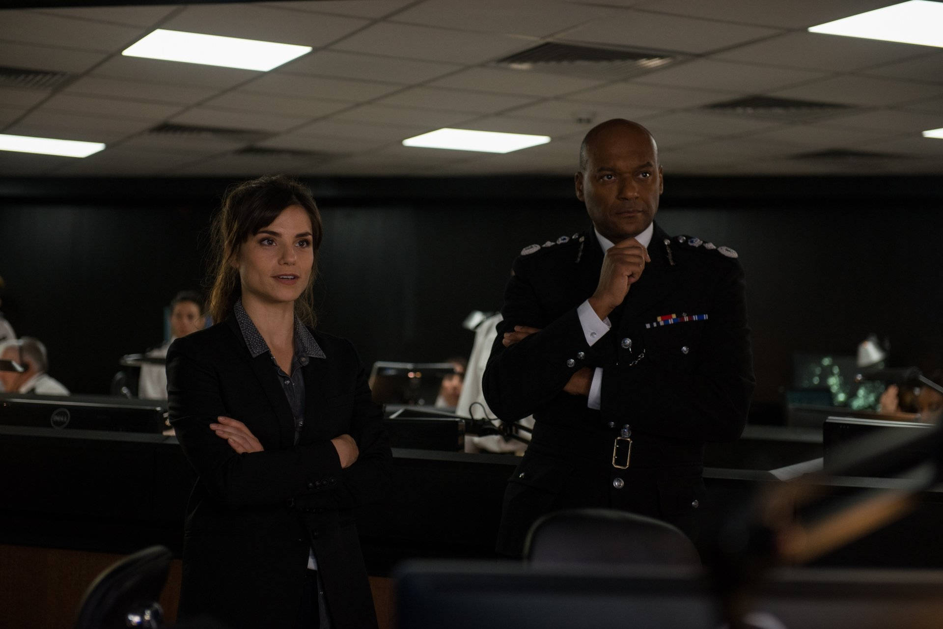 Colin Salmon 桌布 壁紙 — 兩位嚴肅的專業人士站在燈光昏暗的警察指揮中心，螢光燈下設有顯示器和辦公桌。
