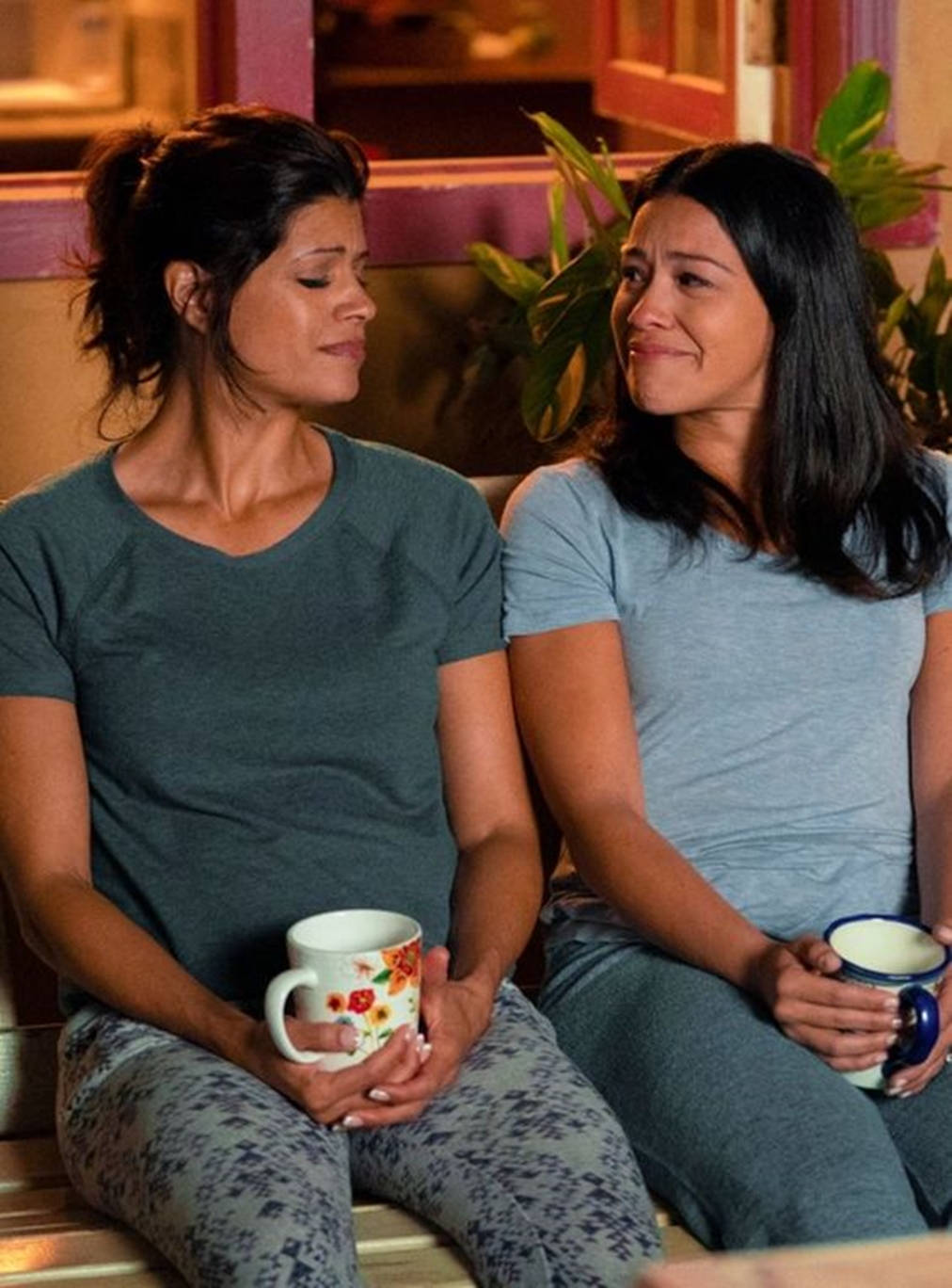 tv 桌布 壁紙 — 兩位女士並排坐在長椅上，手持杯子，在溫暖的燈光和附近的綠色植物下，共享寧靜的室內時光。