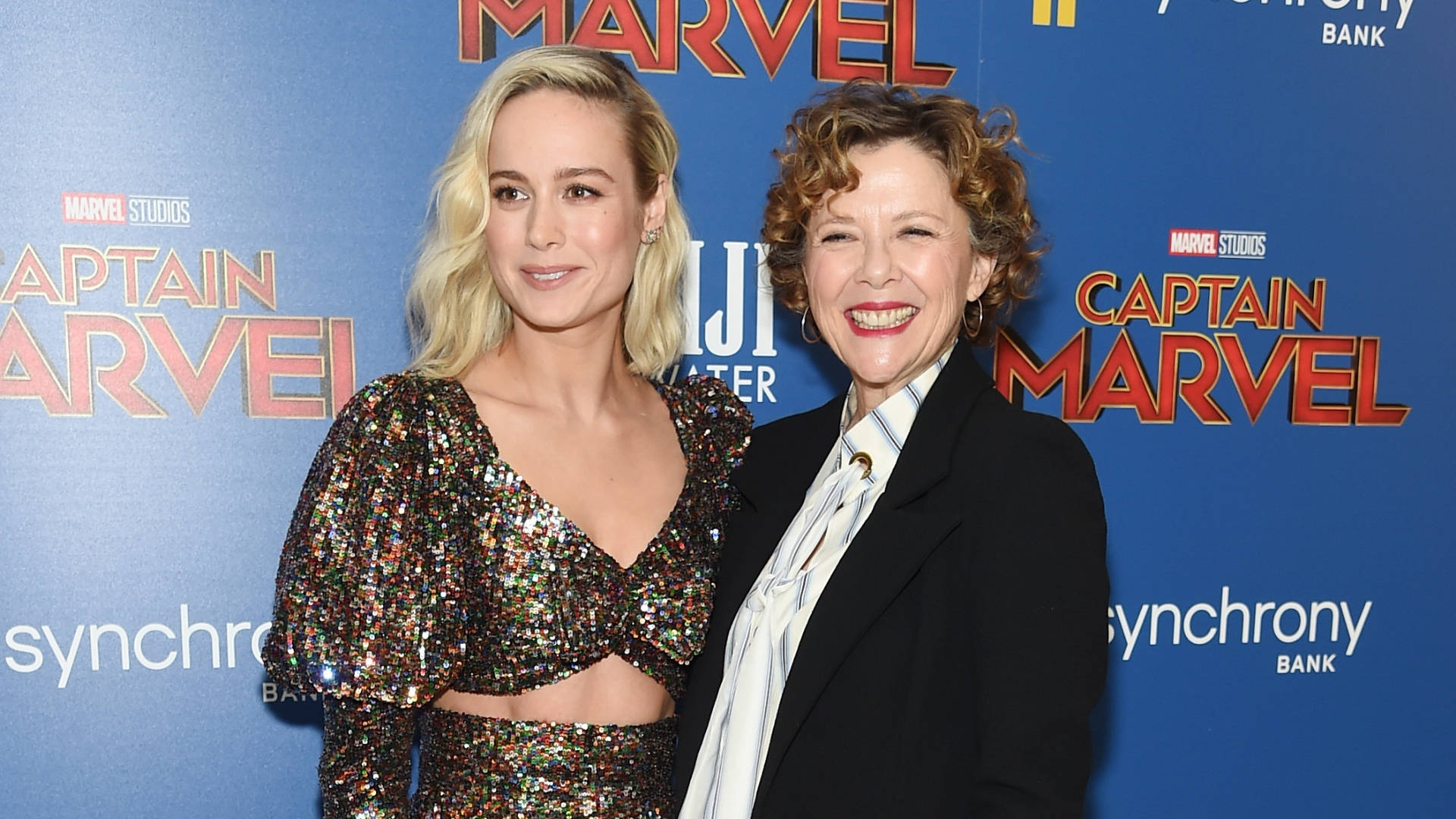 Celebrity 桌布 壁紙 — 兩位女士在《驚奇隊長》首映會上微笑，身穿閃亮亮片上衣，背景為藍色品牌佈景。