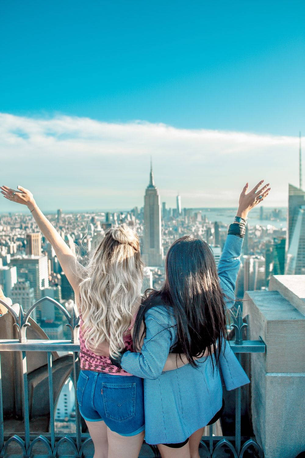 Best Friends 桌布 壁紙 — 兩位女士在一個可俯瞰紐約市標誌性天際線的屋頂上，高舉雙臂擁抱，背景是明亮的藍天。