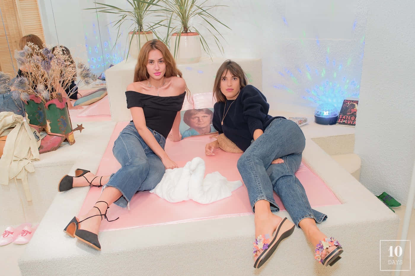 Photoshoot 桌布 壁紙 — 兩位女士在一個明亮、時尚的室內空間中，隨意地躺在白色平台上的粉紅色墊子上，周圍有植物和裝飾品。