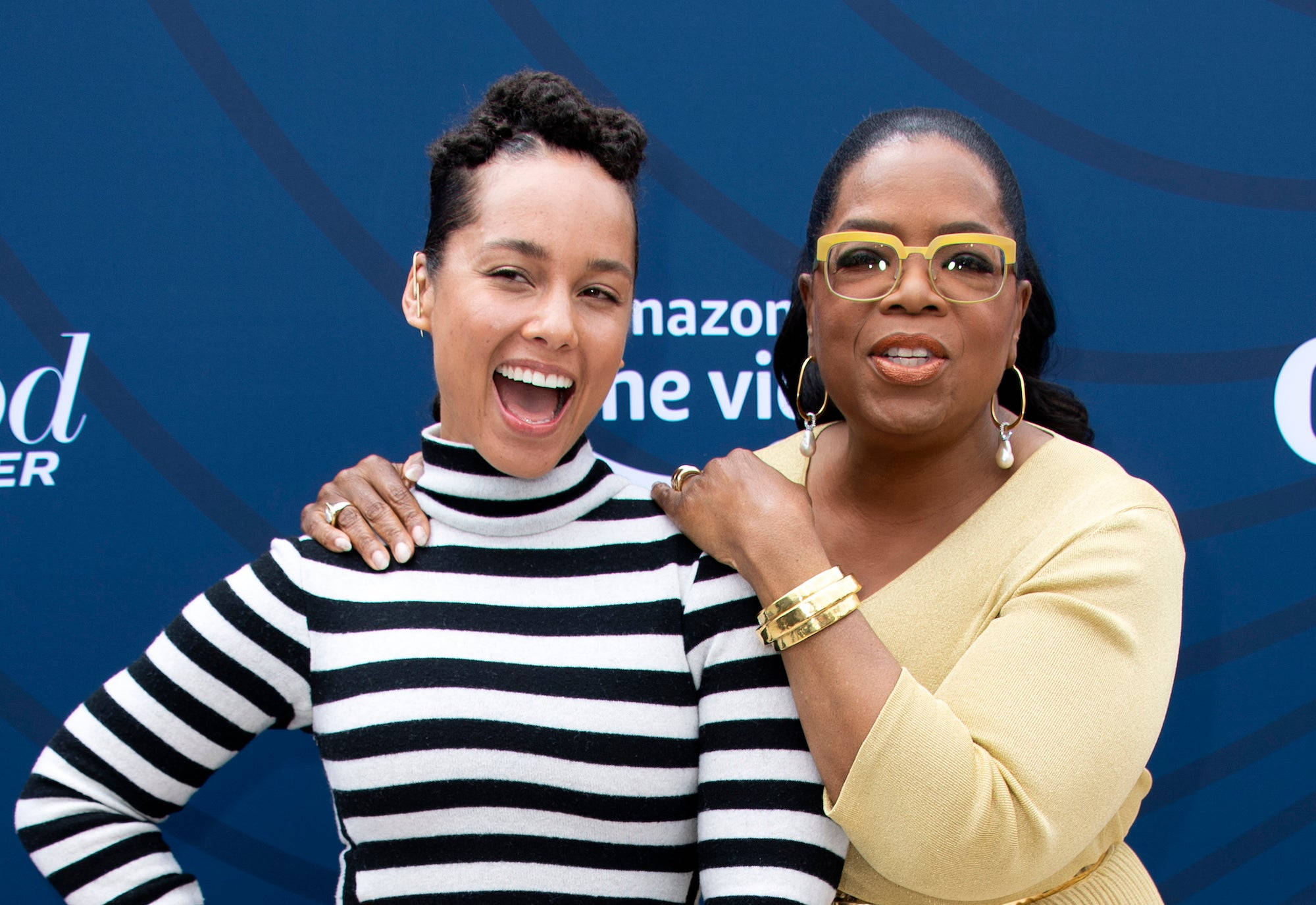 Oprah 桌布 壁紙 — 兩位女士在亞馬遜活動中愉快地擺姿勢，一位穿著條紋毛衣，另一位穿著黃色上衣並戴著大膽的眼鏡。